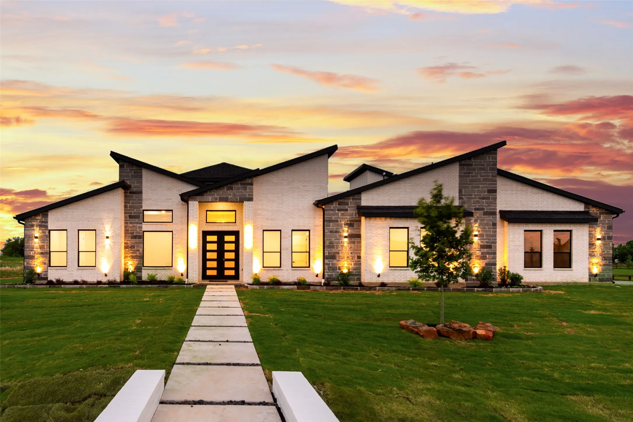 Modern architectural residence featuring a prominent entryway with a multi-pane door, large windows, exterior up-lighting, and a segmented concrete walkway