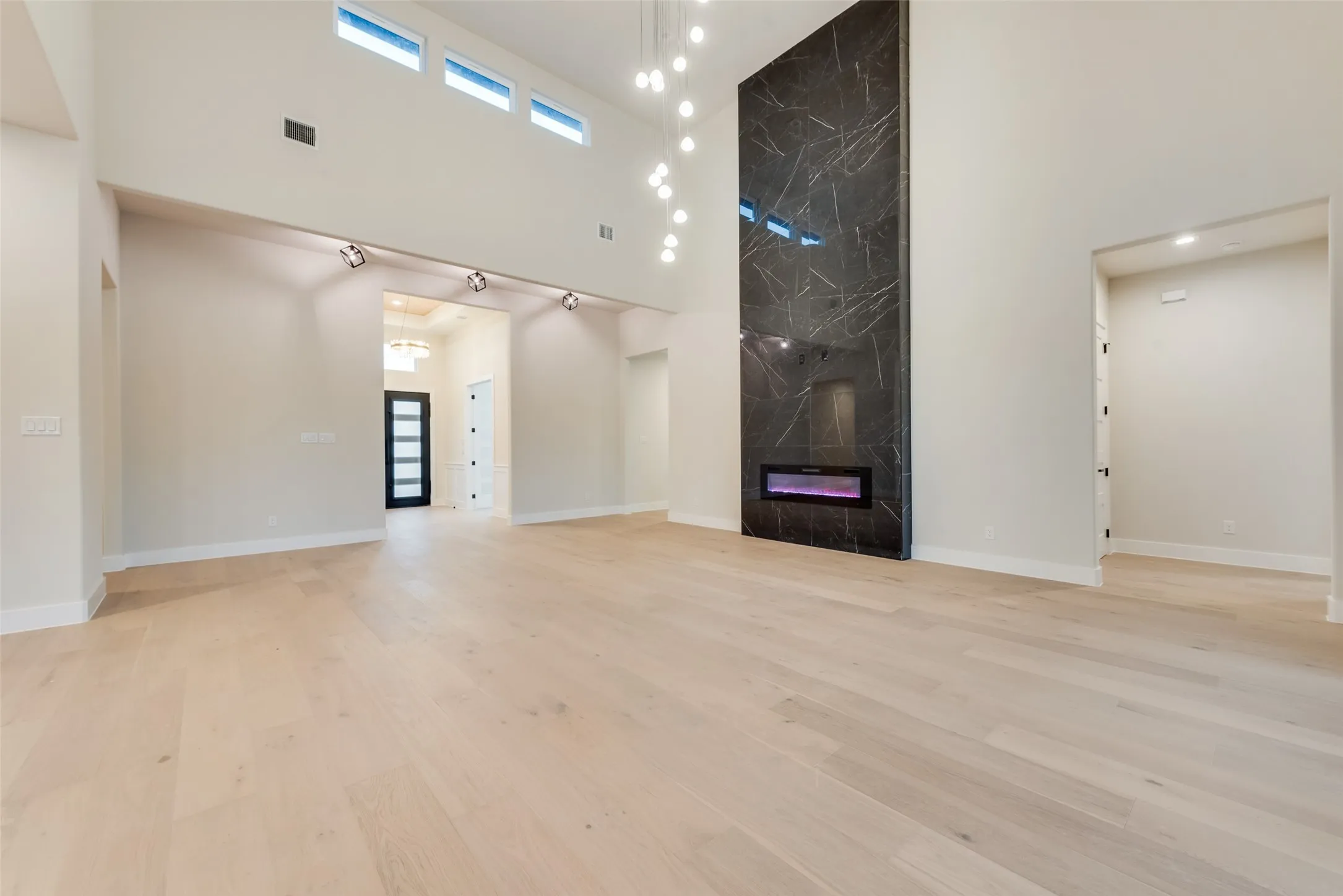 Spacious living area featuring light wood-finish flooring, a tall ceiling with high transom windows, a modern linear electric fireplace with a dark stone-look surround, and a contemporary multi-pendant light fixture