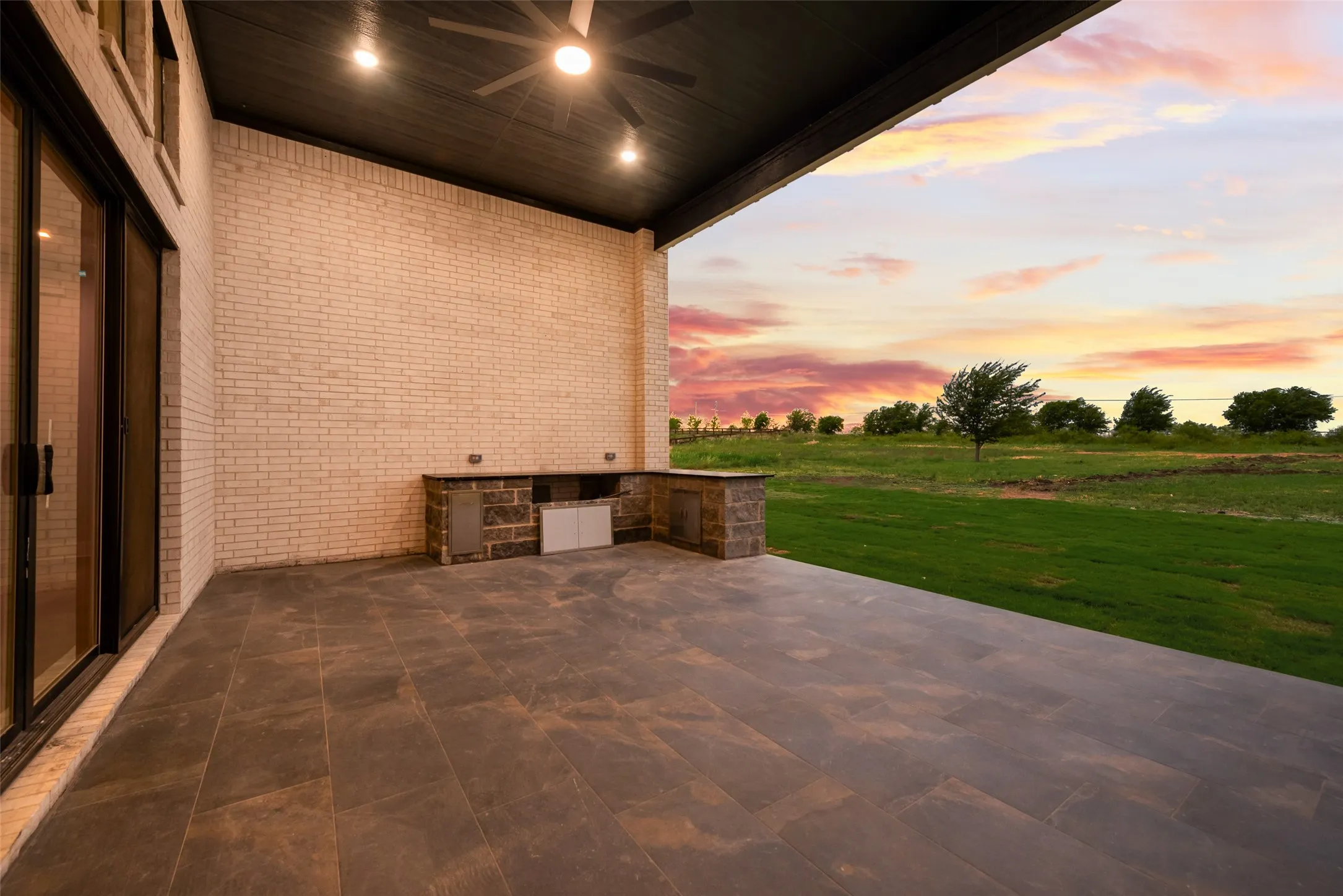 Expansive covered patio featuring large format tile flooring, a built-in outdoor kitchen with stone veneer, and a light-toned brick exterior