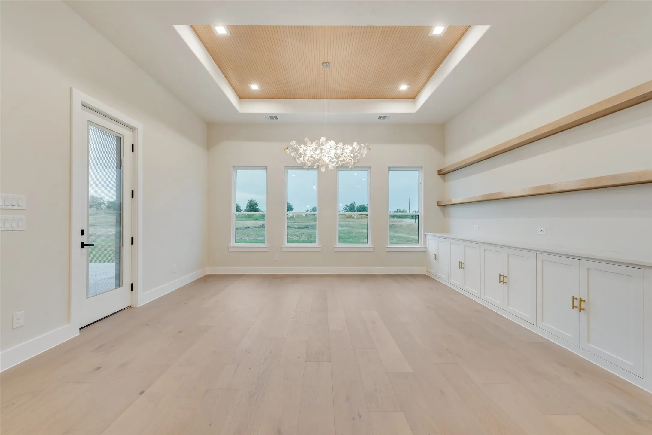 Spacious room featuring light wood-finish flooring, a contemporary chandelier, a coffered ceiling with wood-paneled detail, built-in cabinetry with gold-tone hardware, and three windows offering exterior views