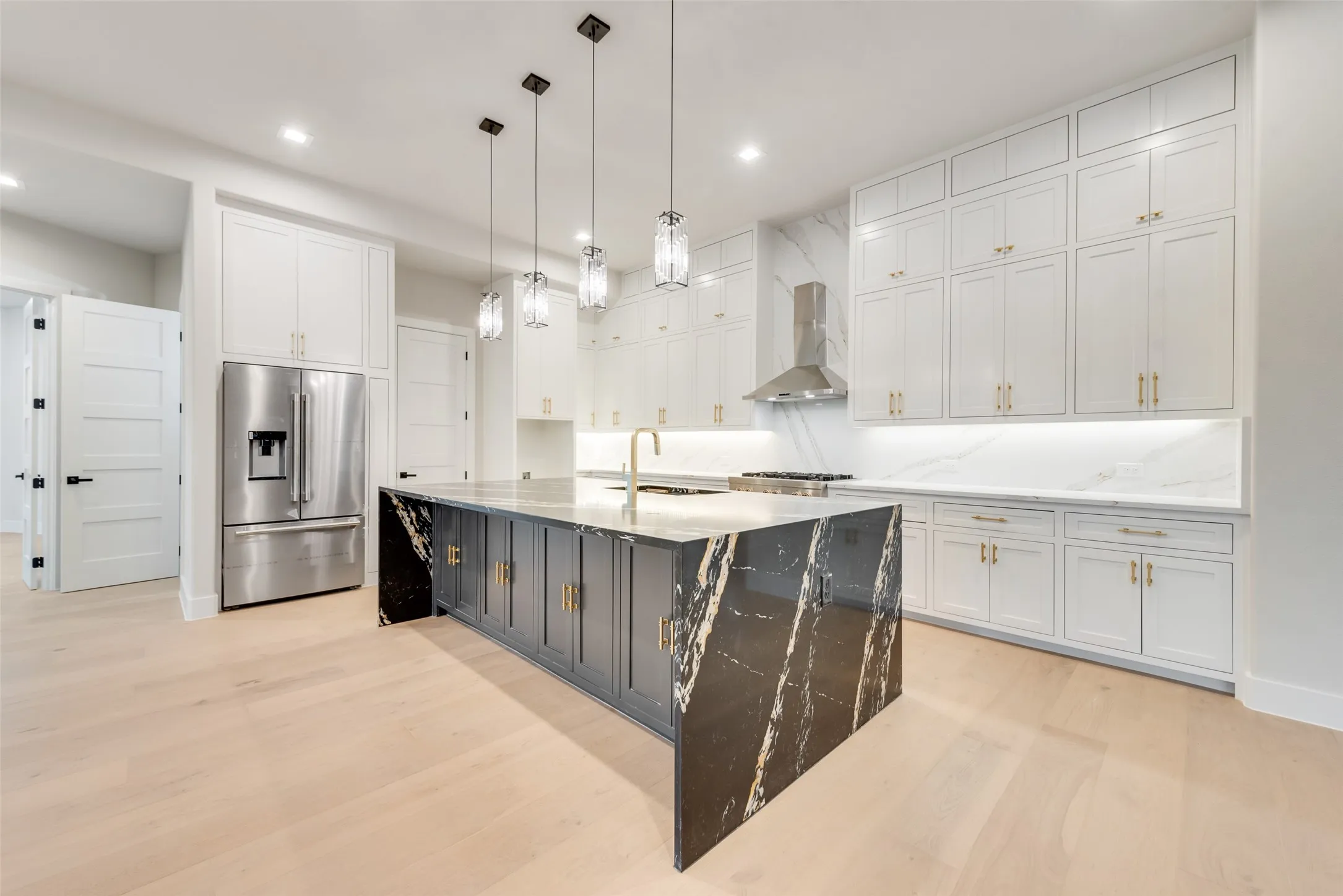 Sleek kitchen featuring a large island with black veined stone, light wood-finish flooring, white cabinetry, integrated stainless steel appliances, and pendant lighting