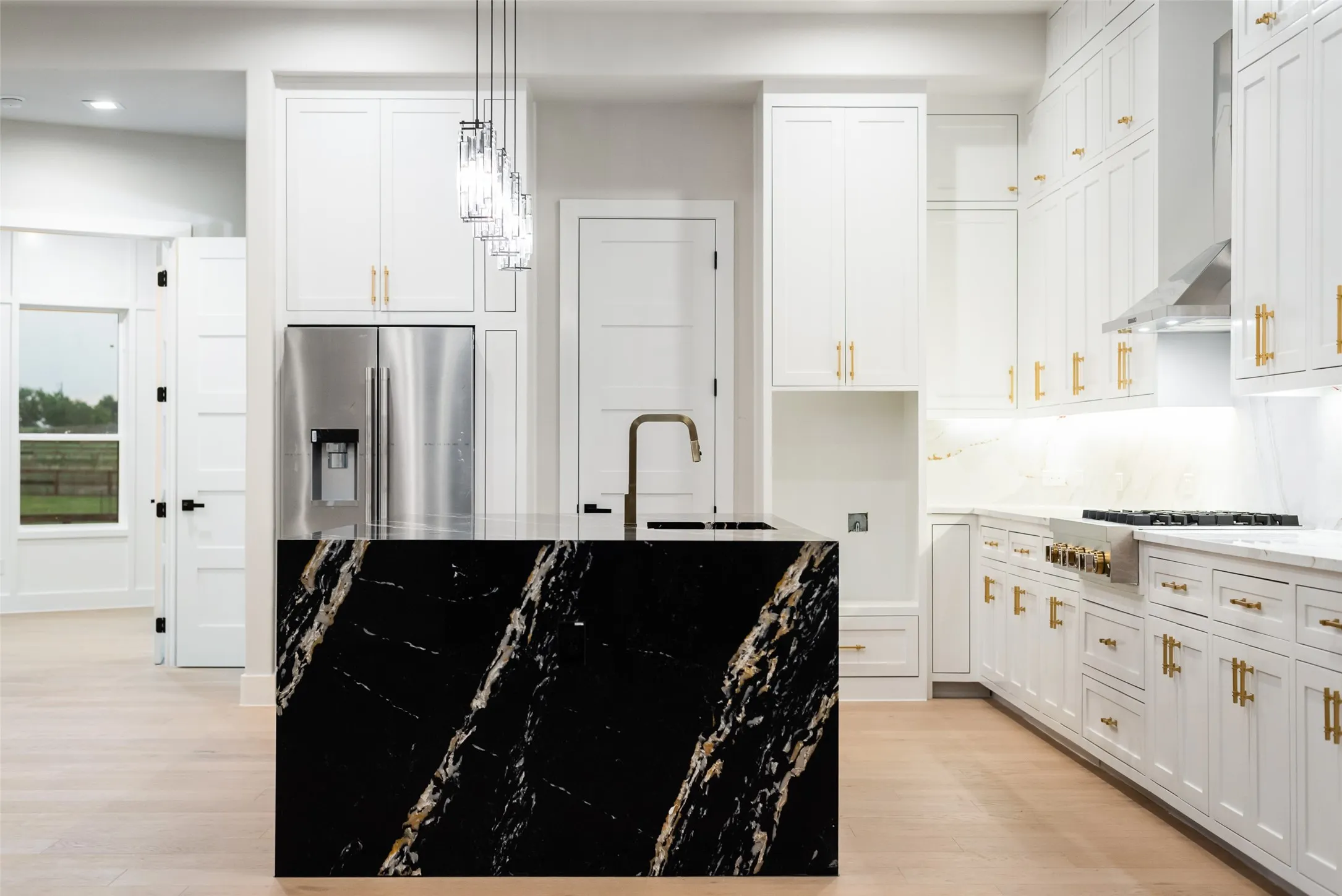 Gourmet kitchen featuring a waterfall island with black stone and gold veining, white shaker cabinetry with gold hardware, light wood-finish flooring, stainless steel range hood, and pendant lighting