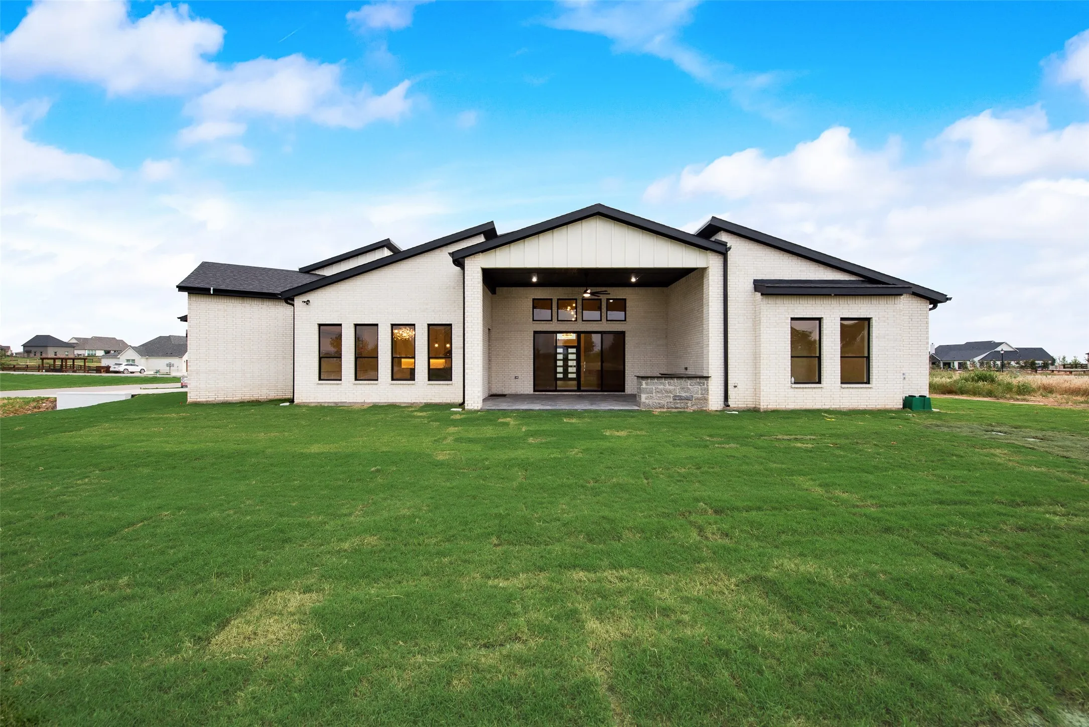 Modern rear exterior featuring a covered patio, extensive grass lawn, sleek black-framed windows, white brick facade, and a built-in outdoor stone counter
