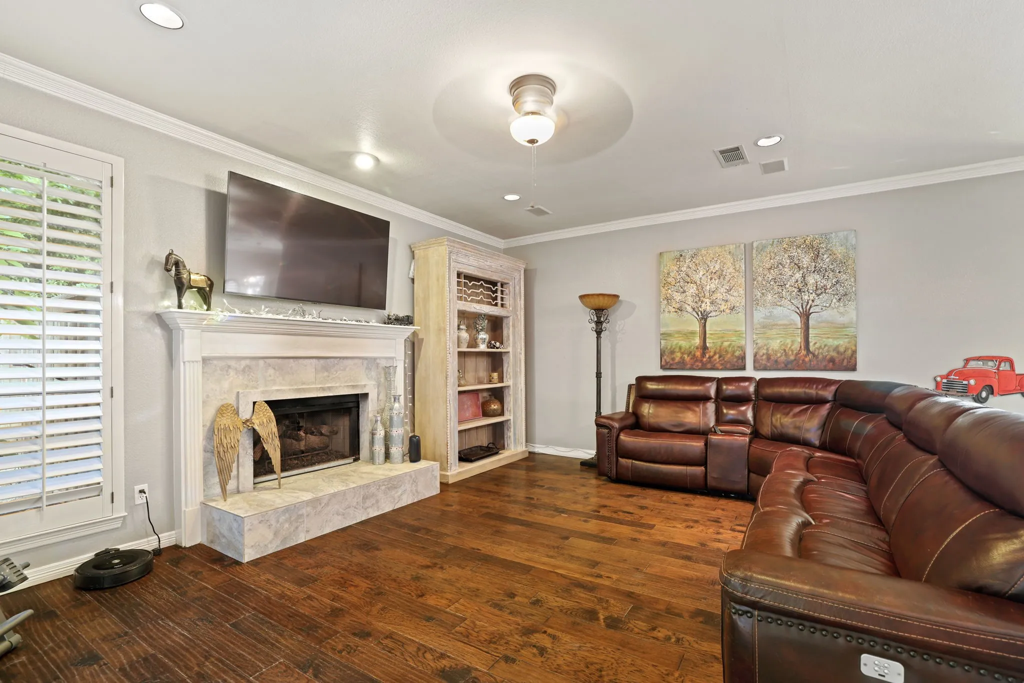 Living area featuring wood-finish flooring, a fireplace with a stone surround and hearth, white plantation shutters, crown molding, and recessed lighting