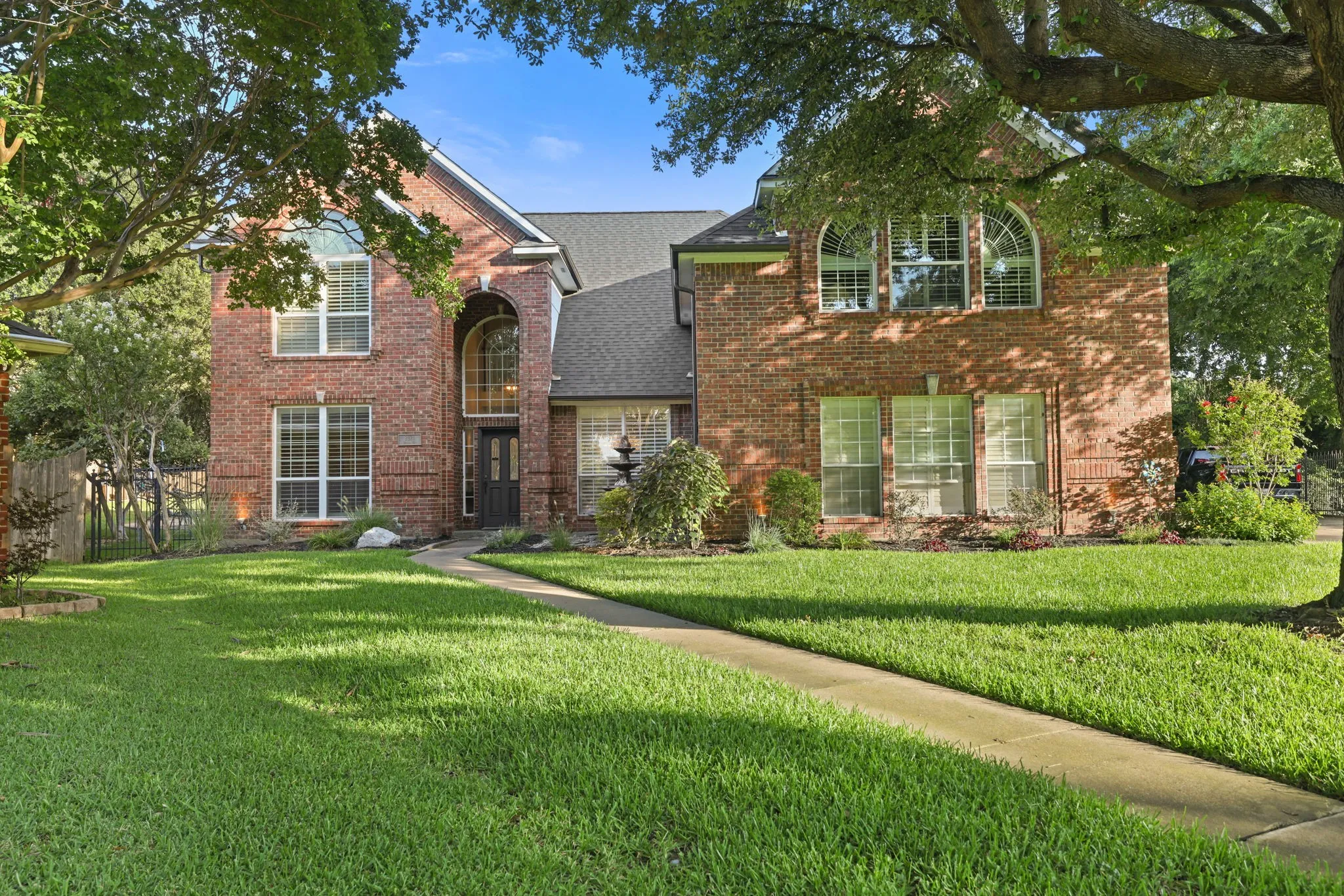 Brick facade residence featuring a front lawn, mature trees, arched entryway, and multiple large windows