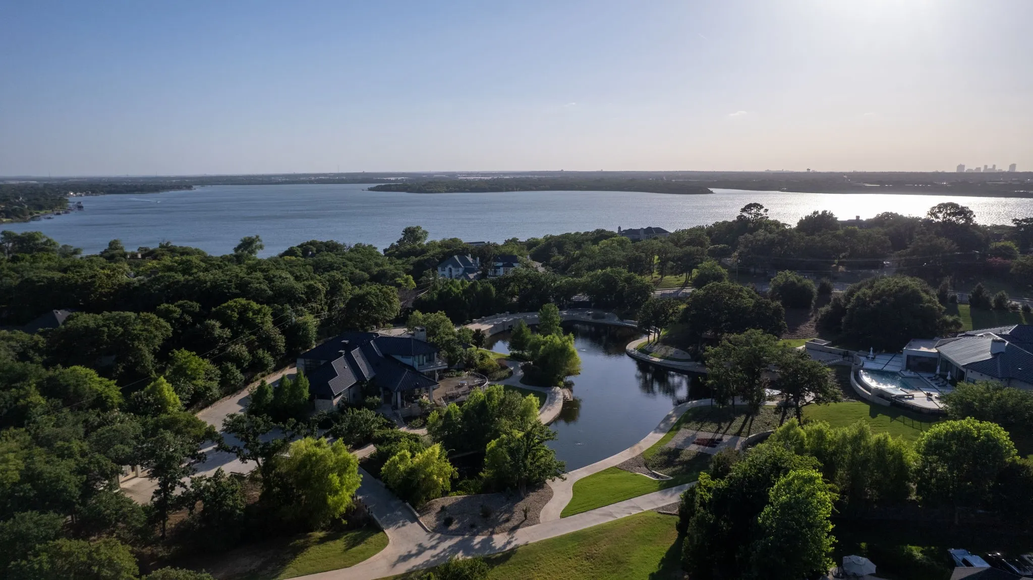 Aerial perspective showcasing a large body of water, lush green tree canopy, winding pathways, and a property featuring a dark-tiled roof and a curved swimming pool