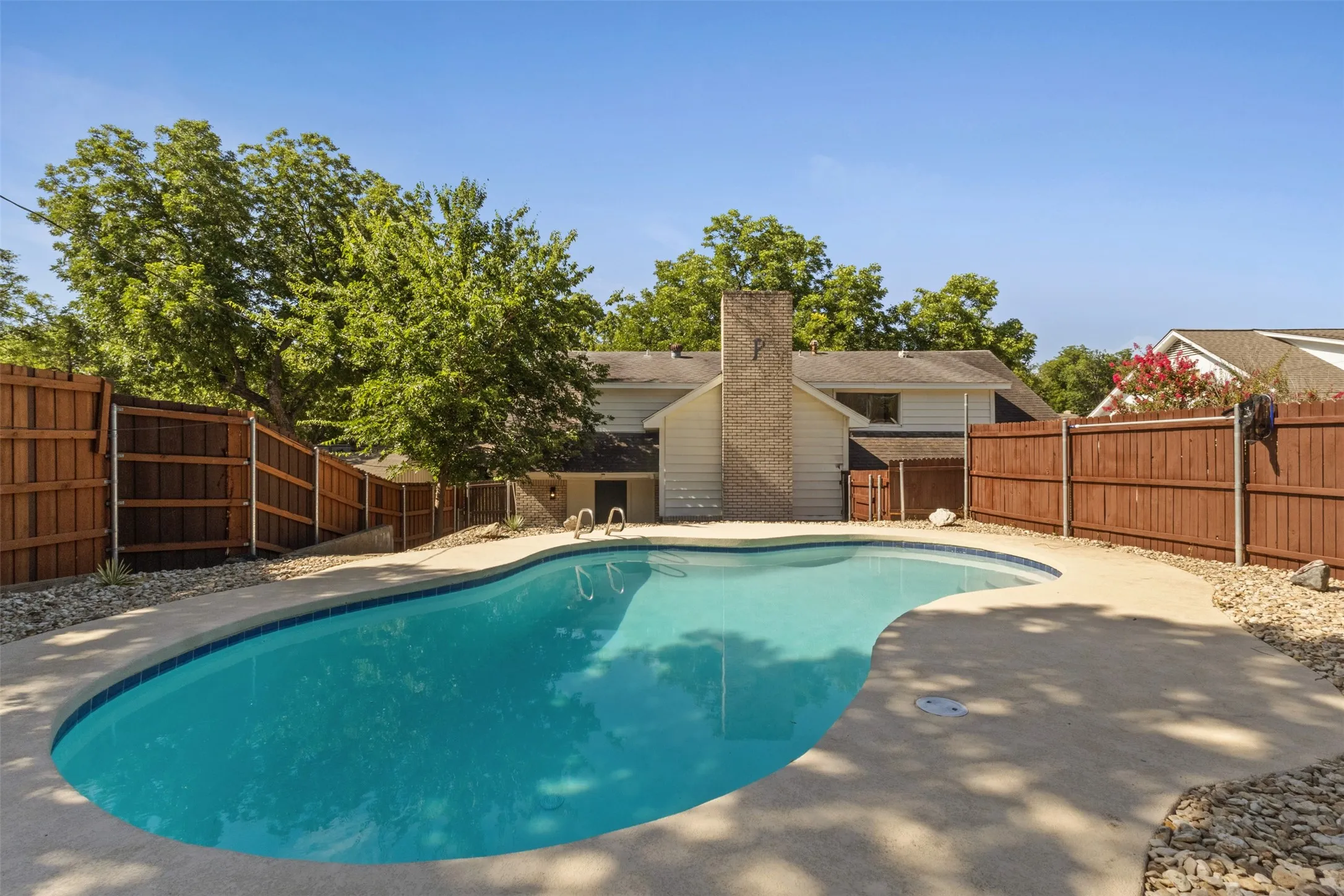 Fenced backyard featuring a freeform swimming pool, extensive concrete decking, and mature trees
