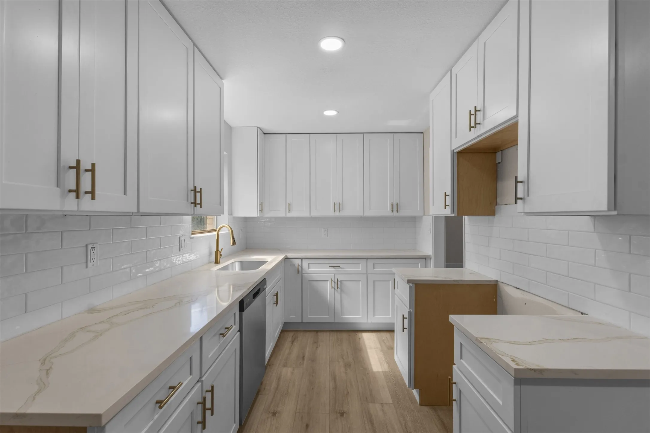 Bright kitchen featuring extensive white shaker cabinetry with gold-tone hardware, light-toned countertops with subtle veining, a white subway tile backsplash, and wood-finish flooring