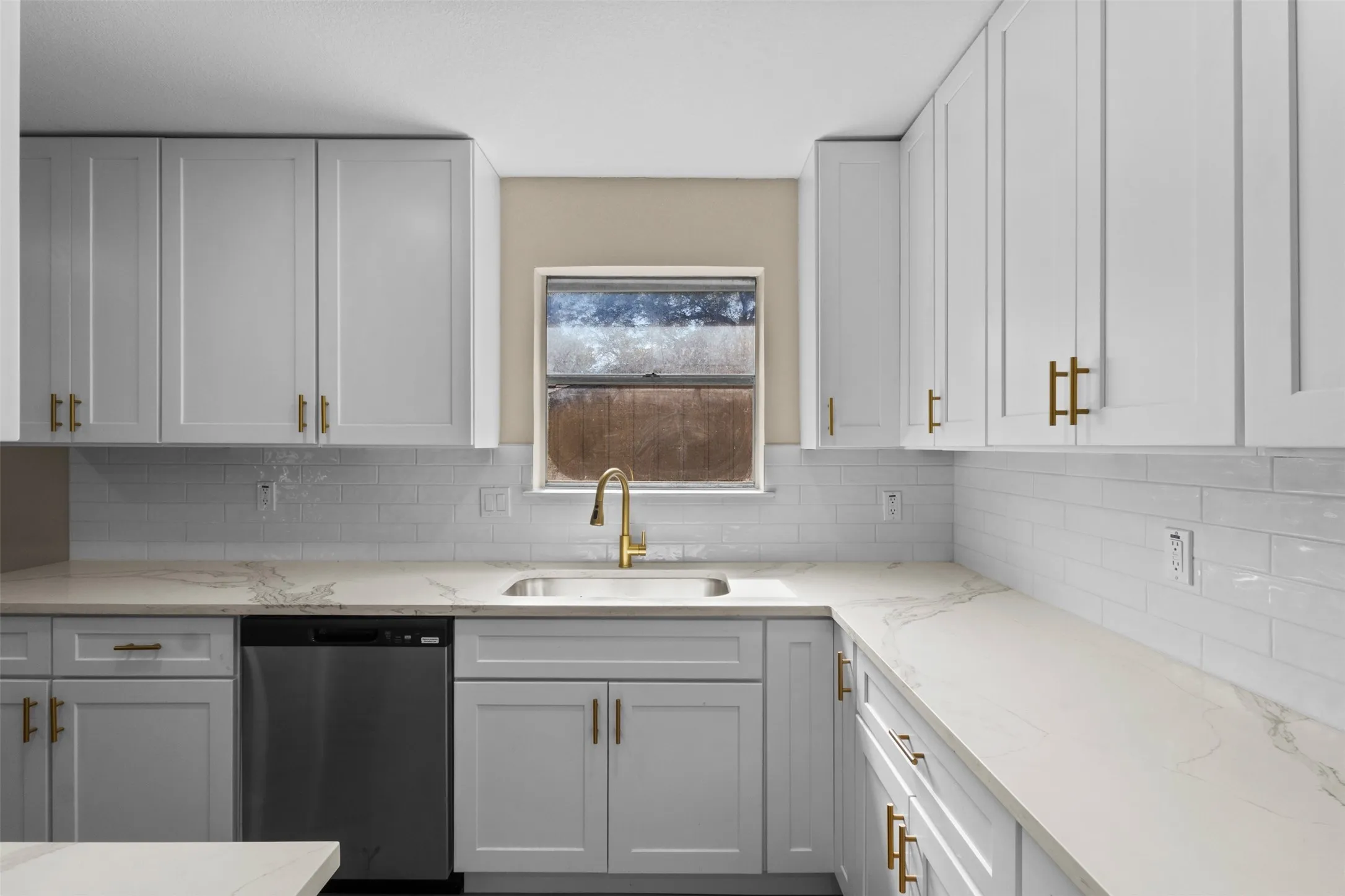 Kitchen featuring white shaker-style cabinetry, gold-finish hardware, light-toned countertops, a stainless steel sink with a gold-finish faucet, and a white subway tile backsplash