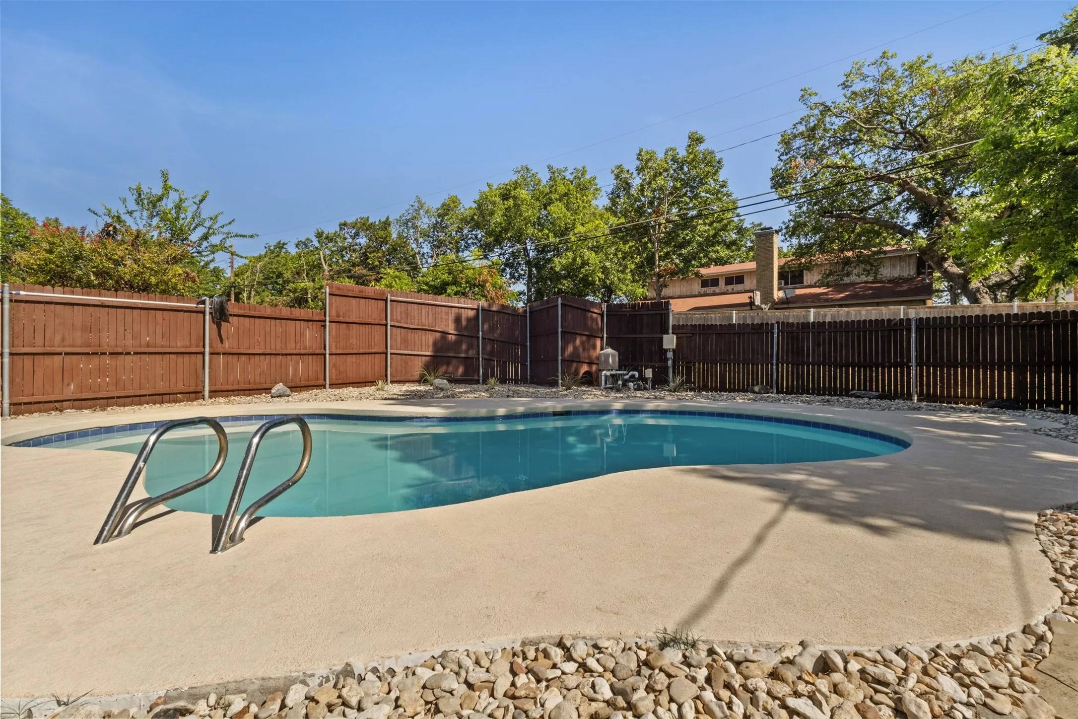 Curved swimming pool featuring a concrete surround, metal handrails, and a perimeter of decorative river rock
