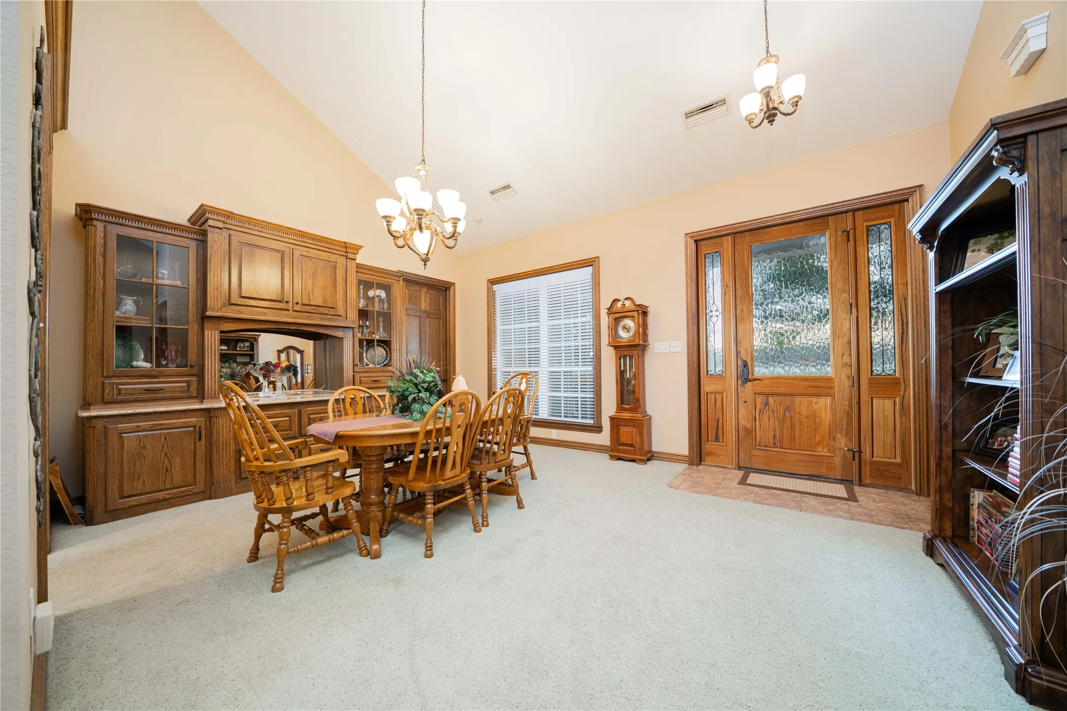 Vaulted ceiling space featuring a large wooden entry door with frosted glass panels, built-in wooden cabinetry with glass-front display cases and a light-colored countertop, overhead chandelier lighting, and light-toned carpet flooring