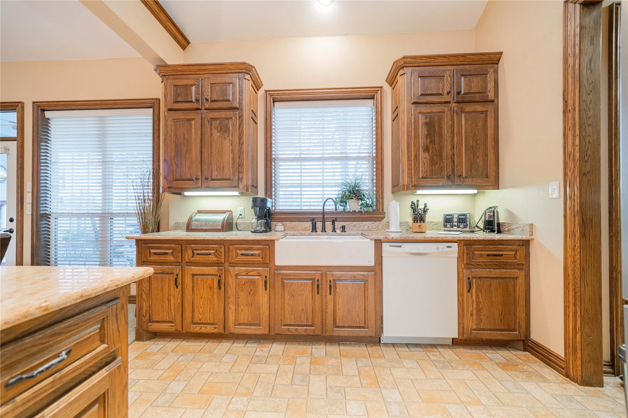 Kitchen featuring natural wood cabinetry, light-toned countertops, a white farmhouse-style sink, and a built-in dishwasher
