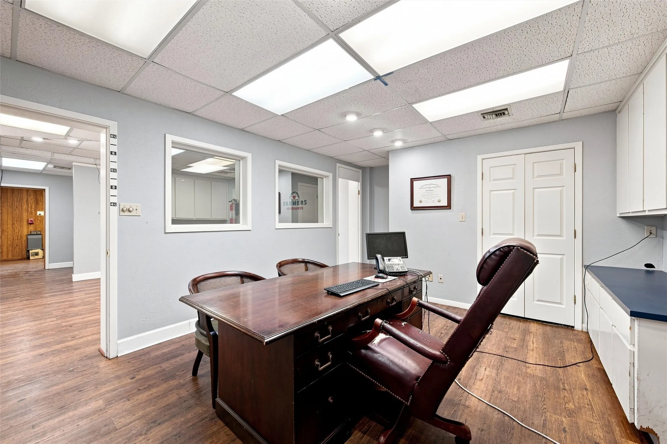 Office space featuring wood-finish flooring, drop ceiling with recessed lighting, light-colored walls, interior windows, and white cabinetry with a dark countertop