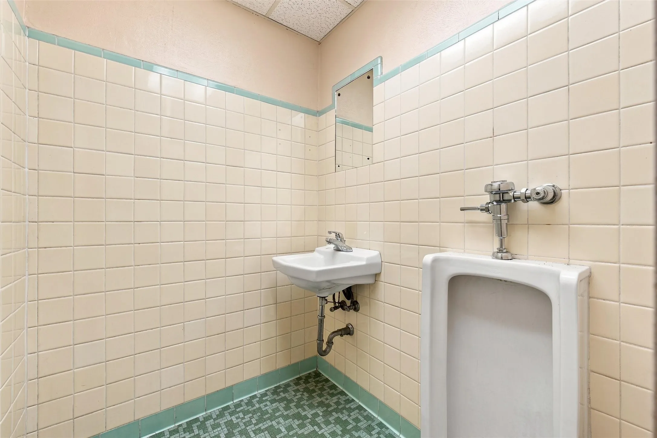 Wall-mounted urinal with chrome flush valve, white wall-mounted sink with chrome faucet, full-height beige square wall tiling, green mosaic floor tiling, and a drop ceiling