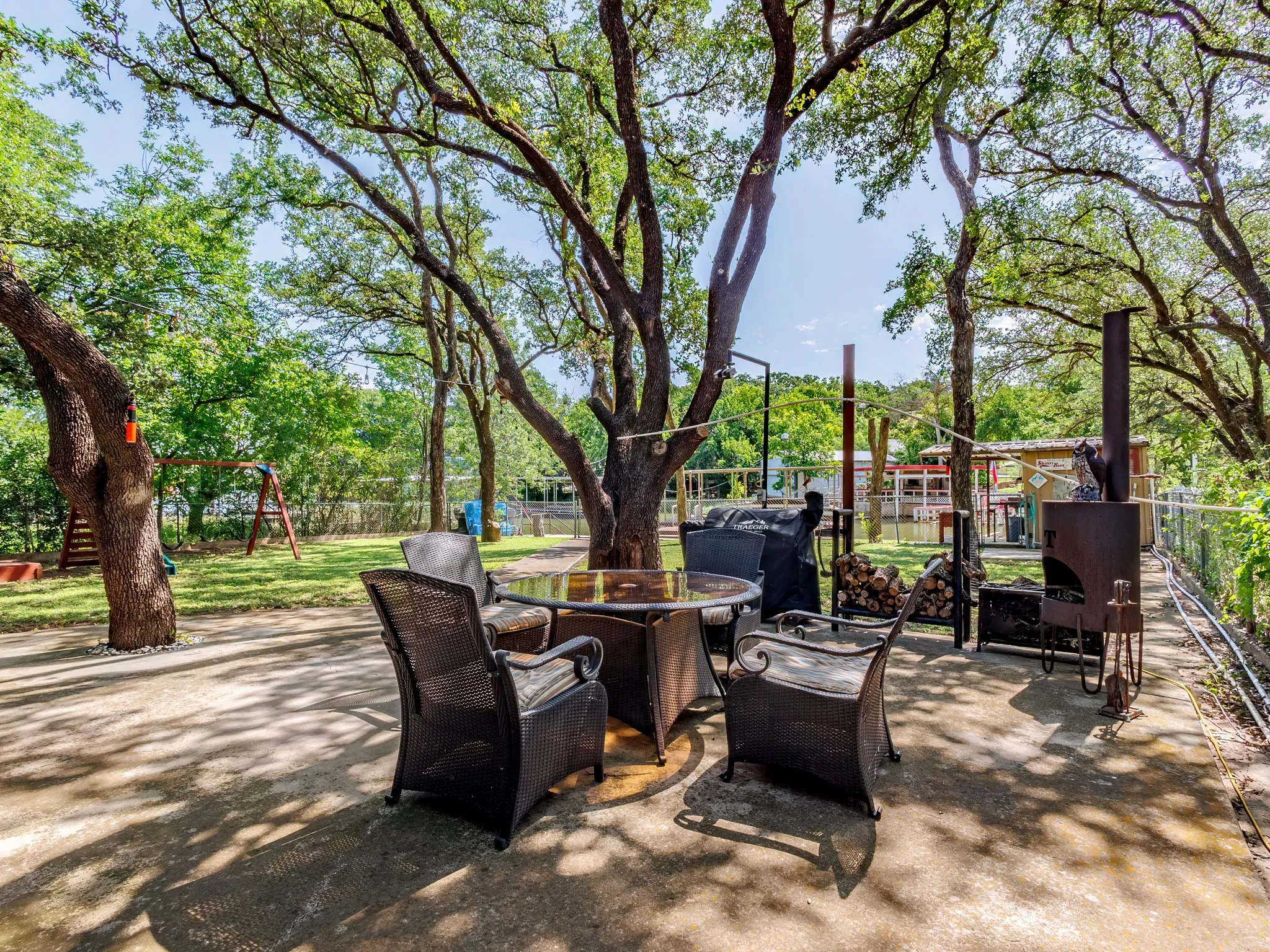 The patio, featuring a grill, table, and chairs beneath mature trees.