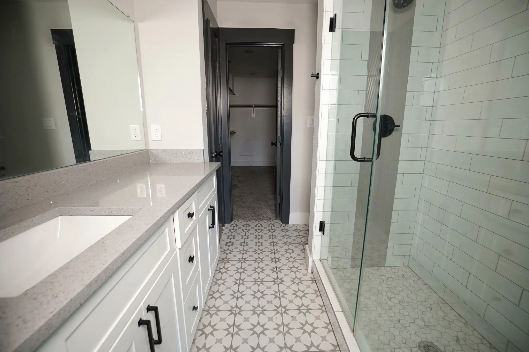 Dual vanity with gray quartz countertop, white cabinetry with black hardware, glass-enclosed shower with subway tile surround, and patterned floor tile