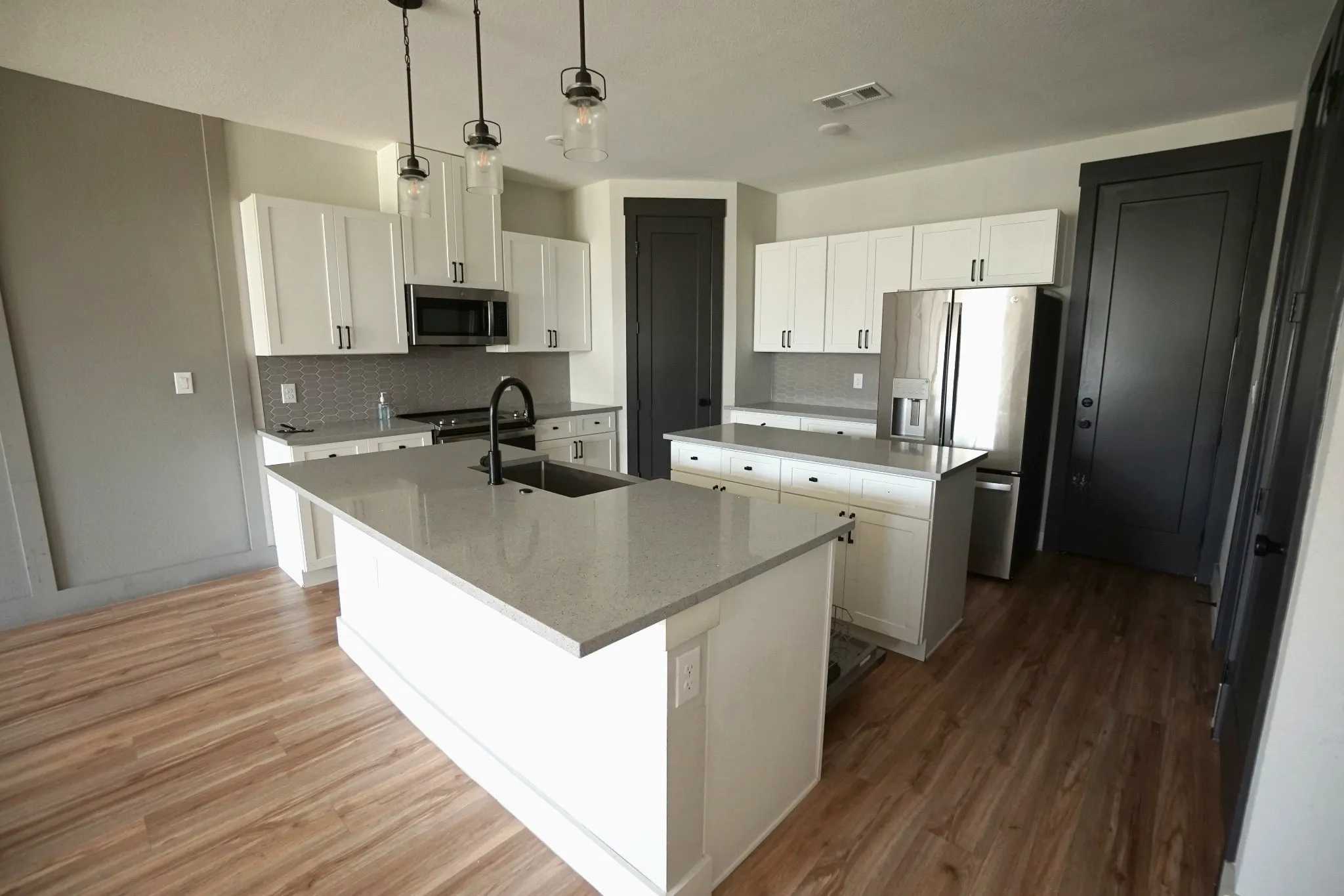 White cabinetry with matte black hardware, stainless steel appliances, wood-finish flooring, island with integrated sink, and pendant lighting