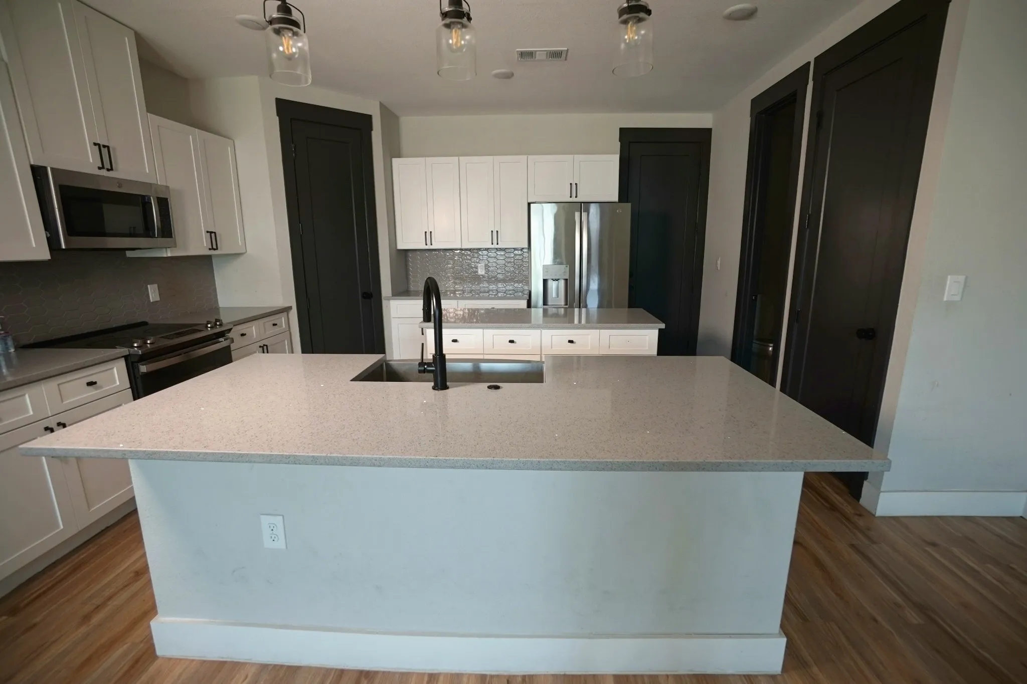 Kitchen featuring an island with an undermount sink, pendant lighting, stainless steel appliances, white cabinetry, and wood-finish flooring