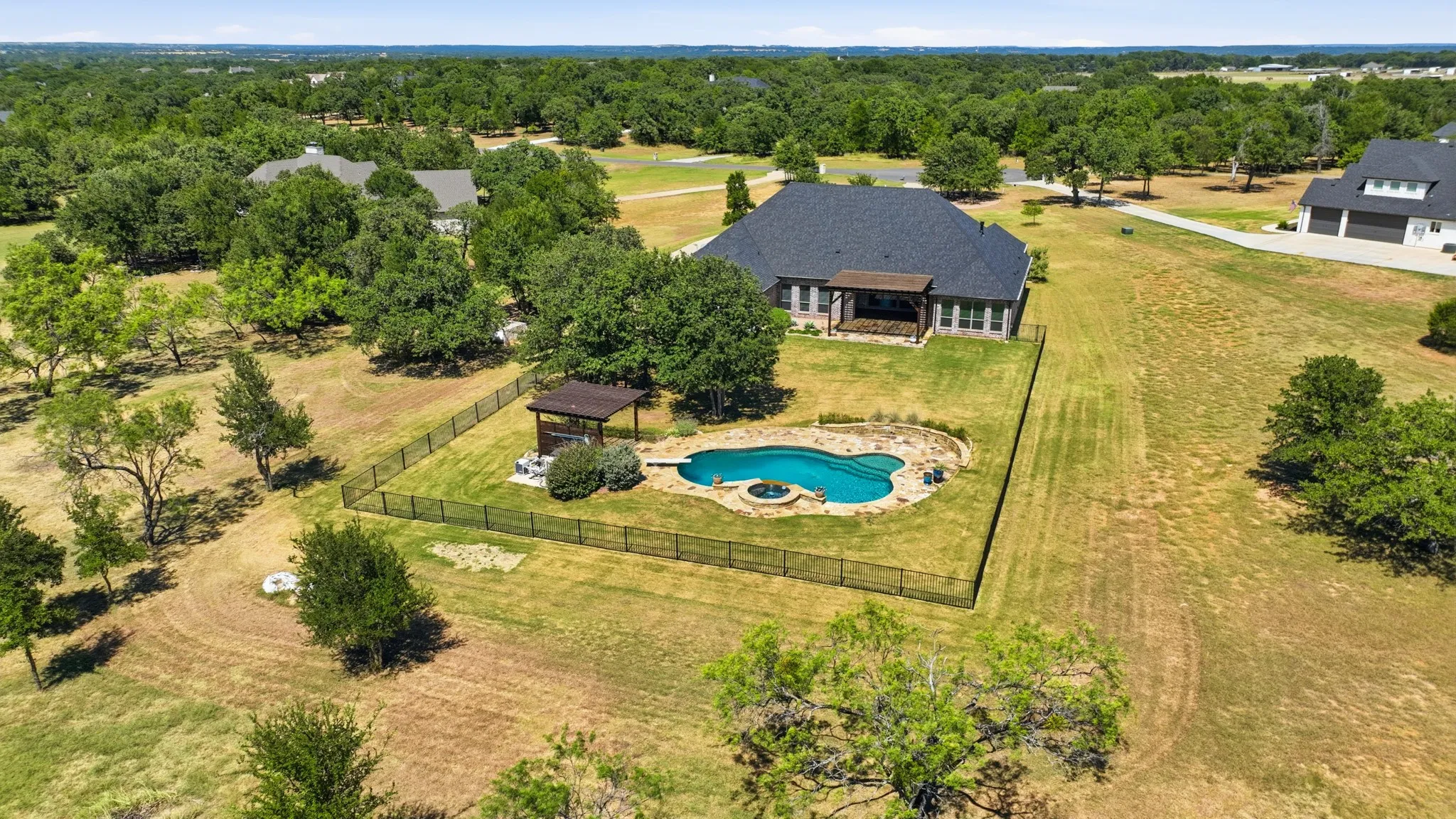 Expansive property featuring a dark-roofed residence, a fenced swimming pool with a water feature, and an adjacent gazebo