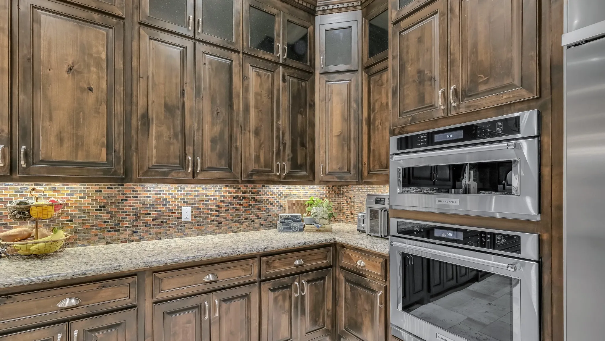 Rich wood cabinetry with varied finishes, stone countertops, and a full-height tile backsplash create a functional kitchen corner