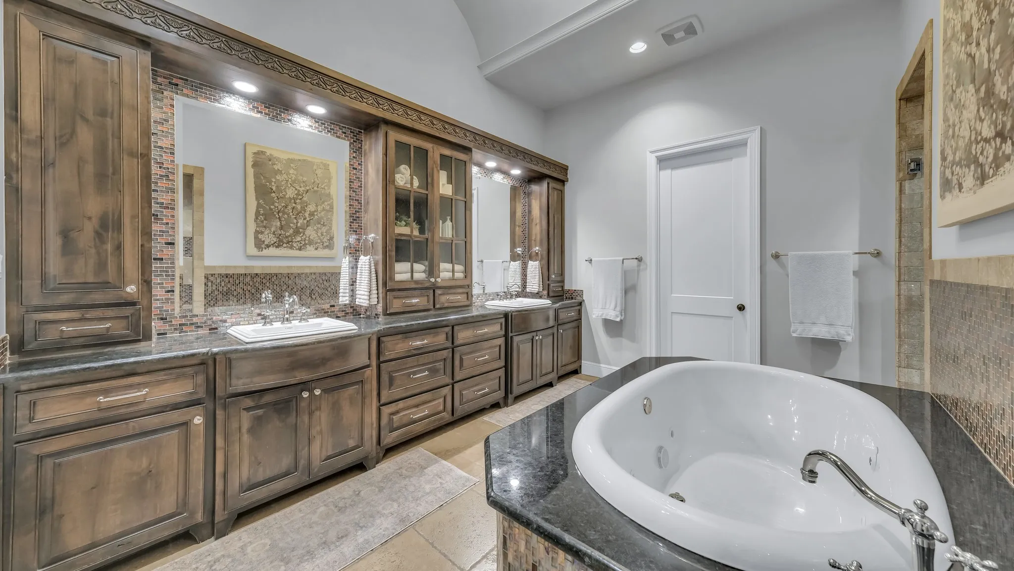 Spacious bathroom featuring a built-in jetted tub, dual wood-finish vanities with undermount sinks, stone countertops, and mosaic tile backsplashes