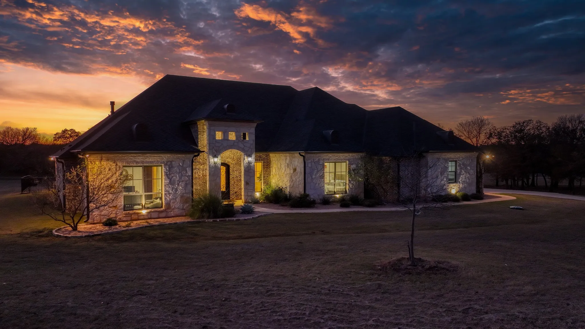 Stone exterior residence featuring a prominent arched entry, a dark shingle roof, and multiple large windows