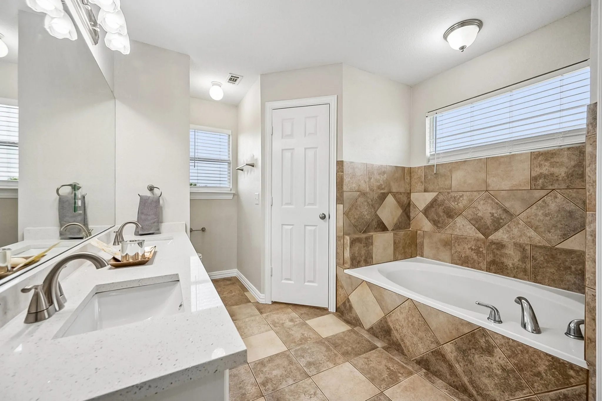 Bathroom featuring a dual vanity with a white countertop and rectangular sinks, brushed nickel fixtures, a large soaking tub with tile surround, and tile flooring