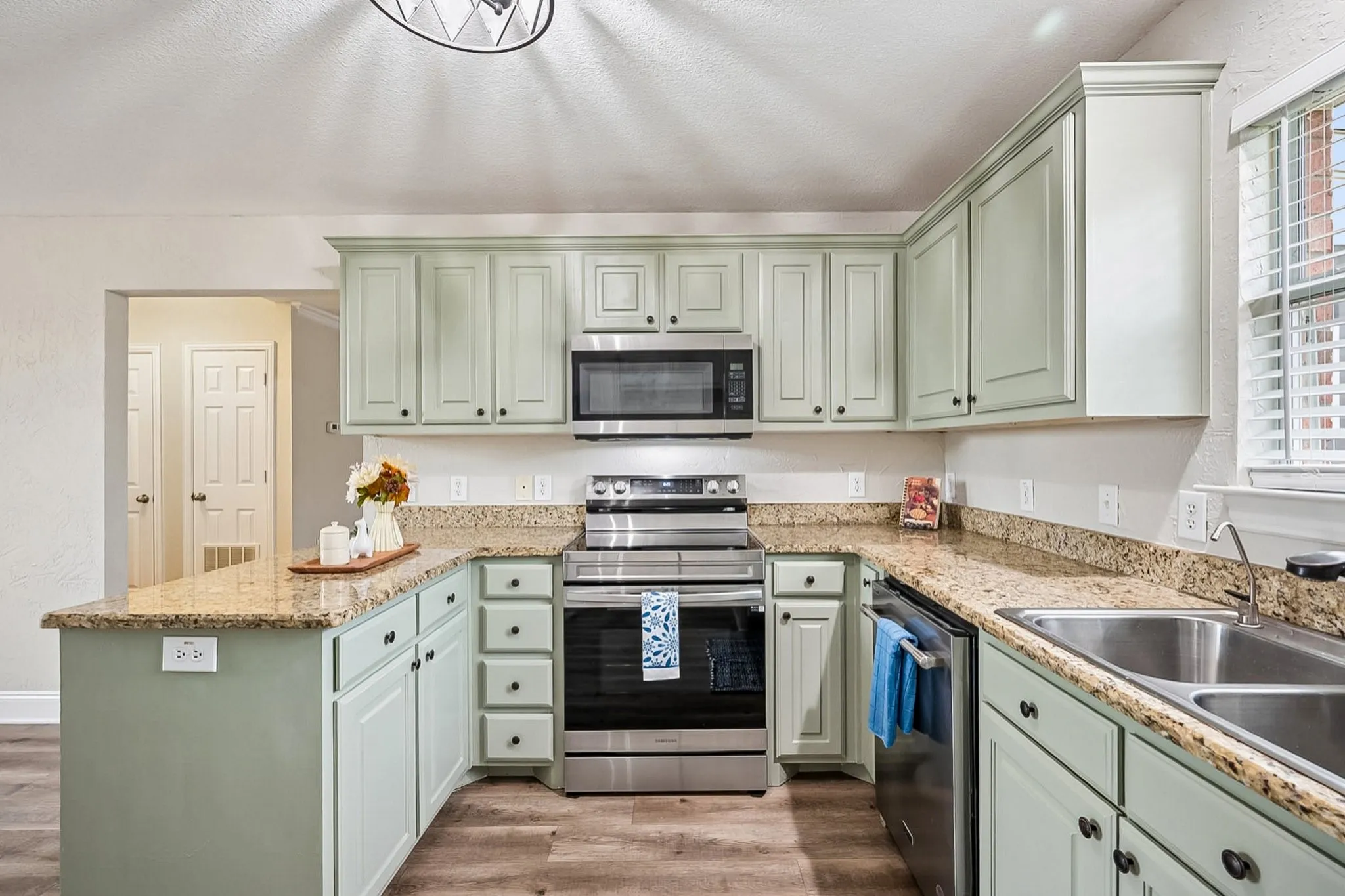 U-shaped kitchen featuring light green cabinetry, granite countertops, stainless steel appliances, a double basin sink, and wood-finish flooring