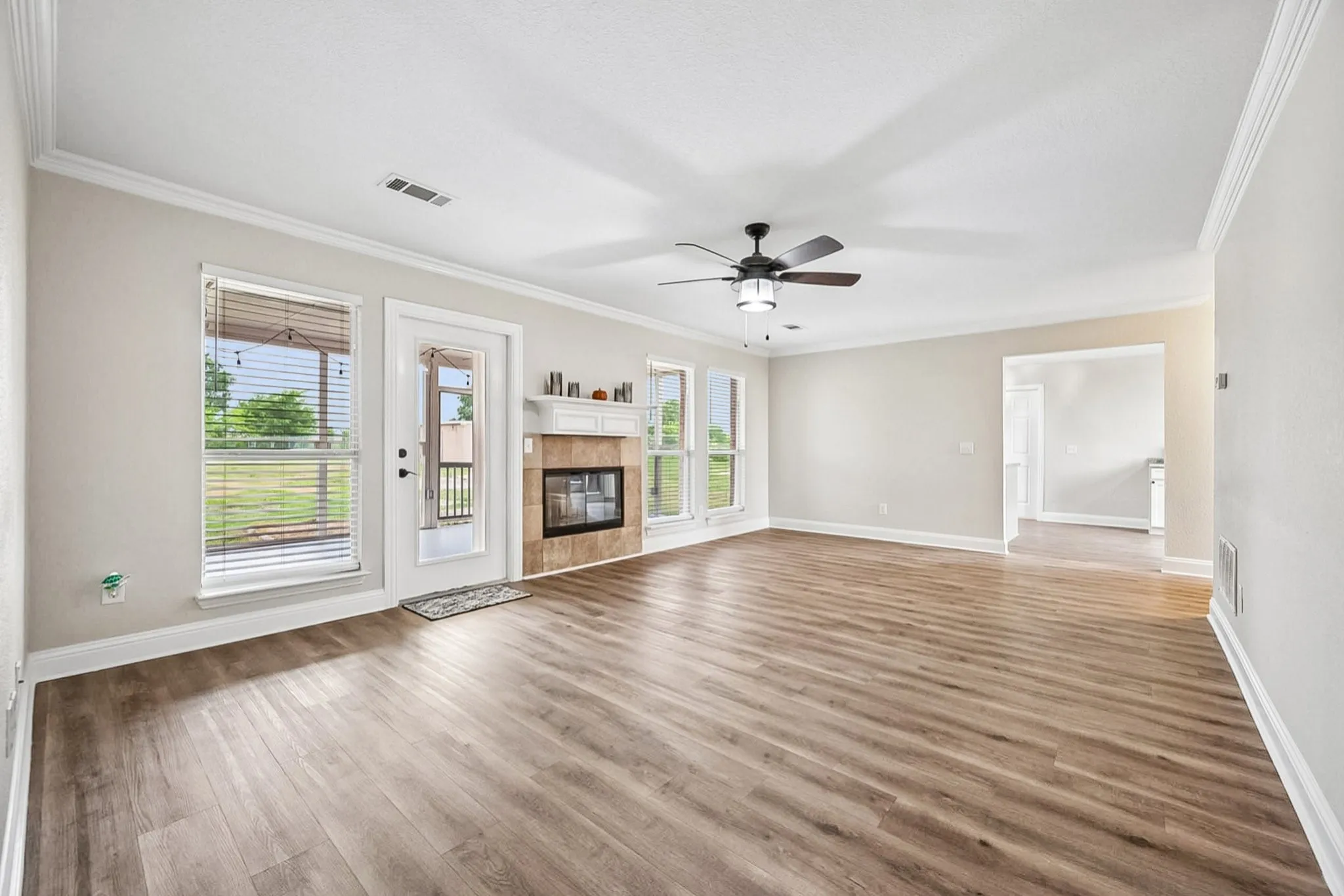 Spacious living area featuring wood-finish flooring, a white mantel fireplace with tile surround, multiple windows, a ceiling fan, and crown molding