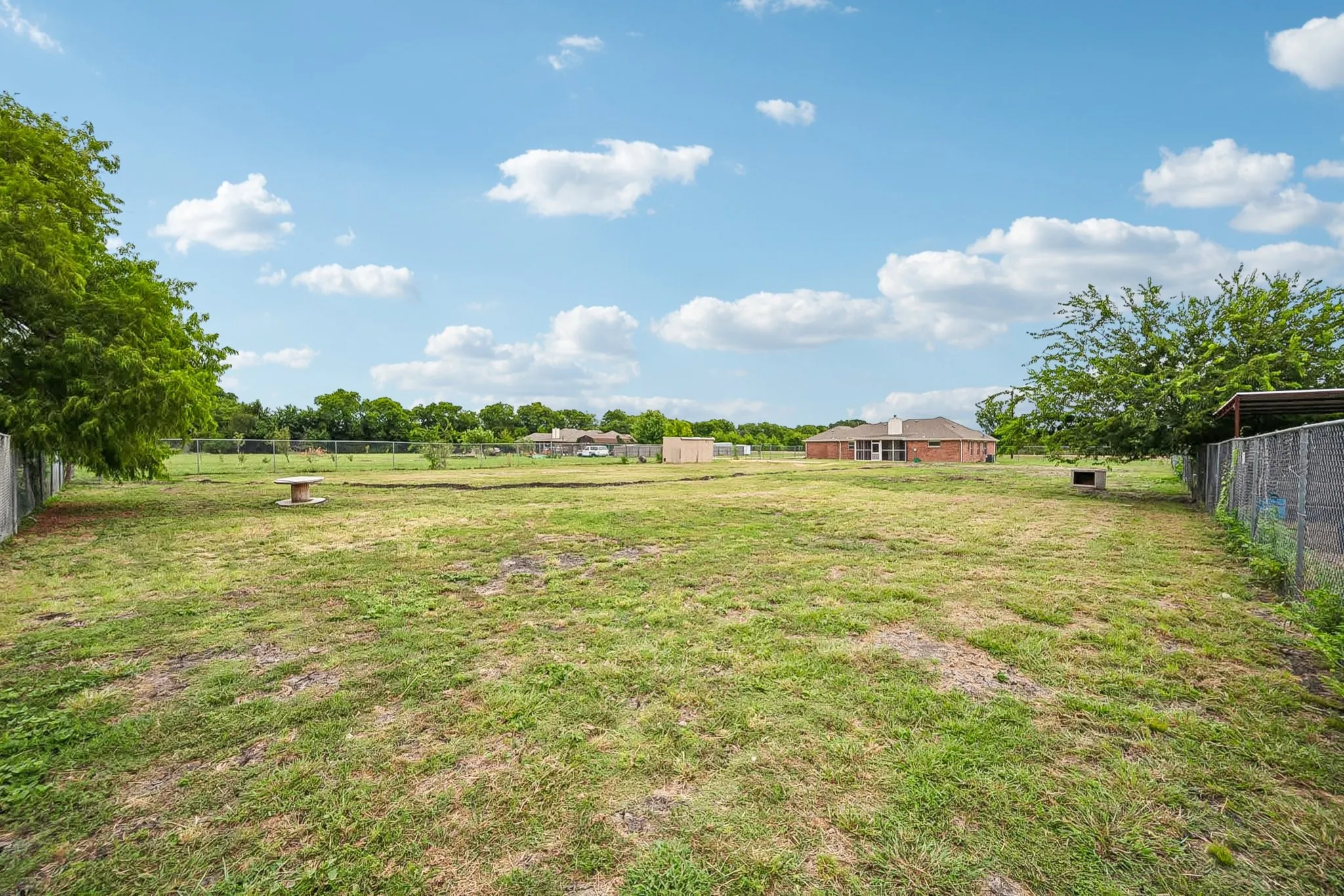 Expansive grassy lot with chain-link fencing, mature trees, and a visible brick residence in the background