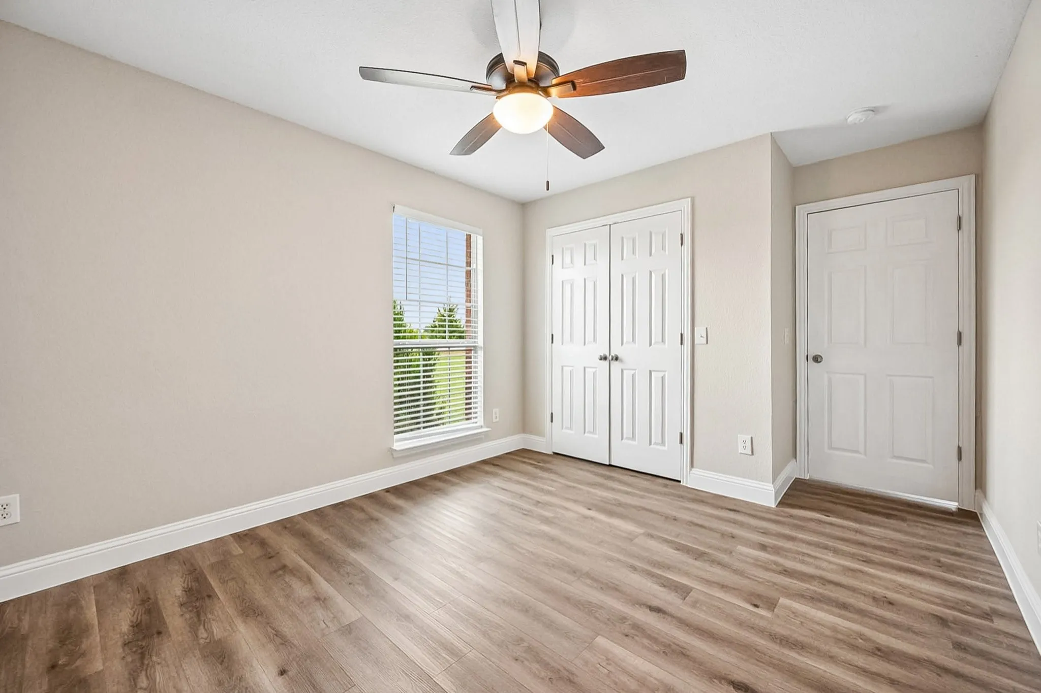 Neutral-toned room featuring wood-finish flooring, a ceiling fan with integrated lighting, a double-door closet, and a single window with horizontal blinds