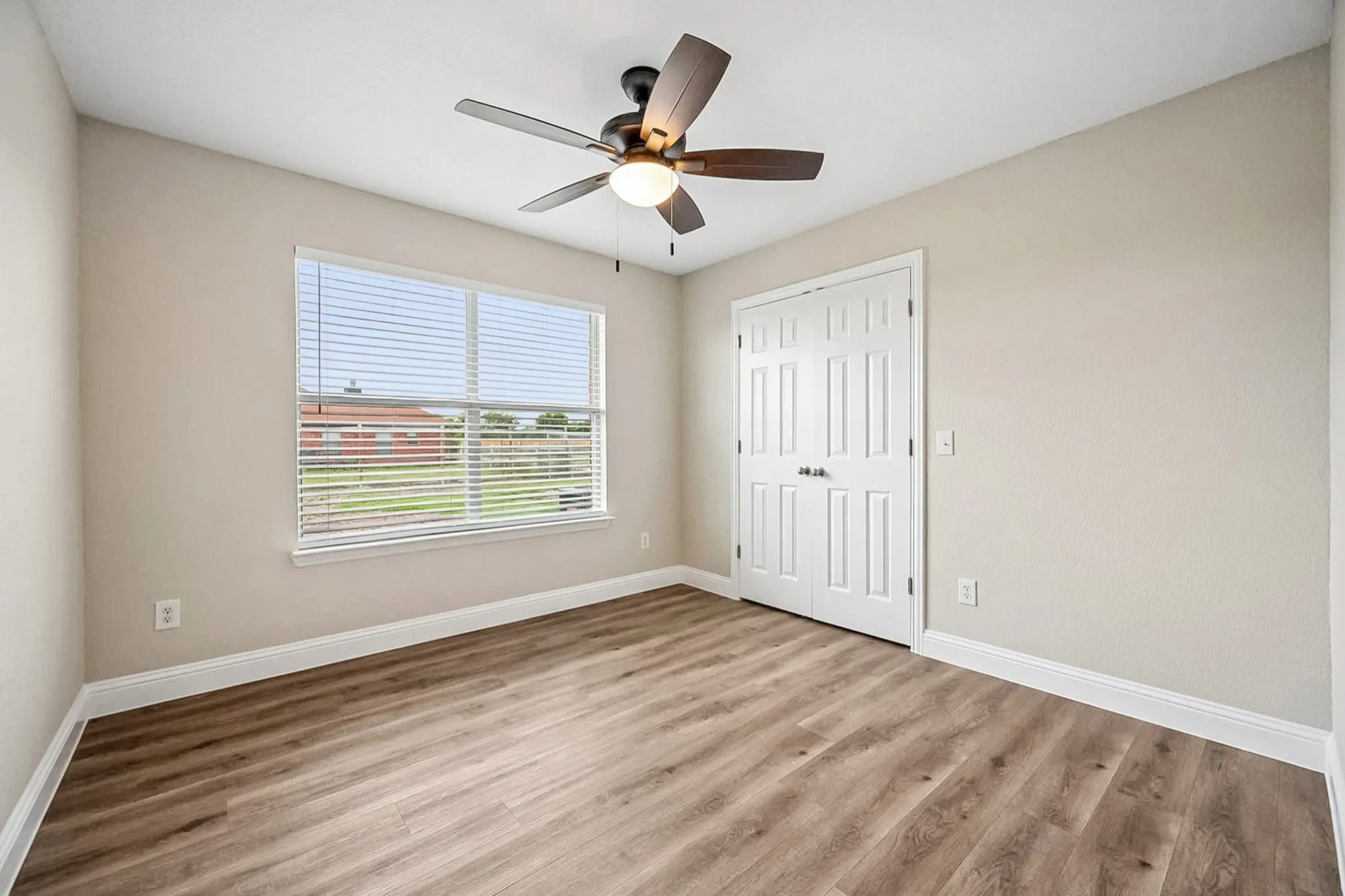 Neutral-toned room featuring wood-finish flooring, a ceiling fan with light, a window with blinds, and white bi-fold closet doors