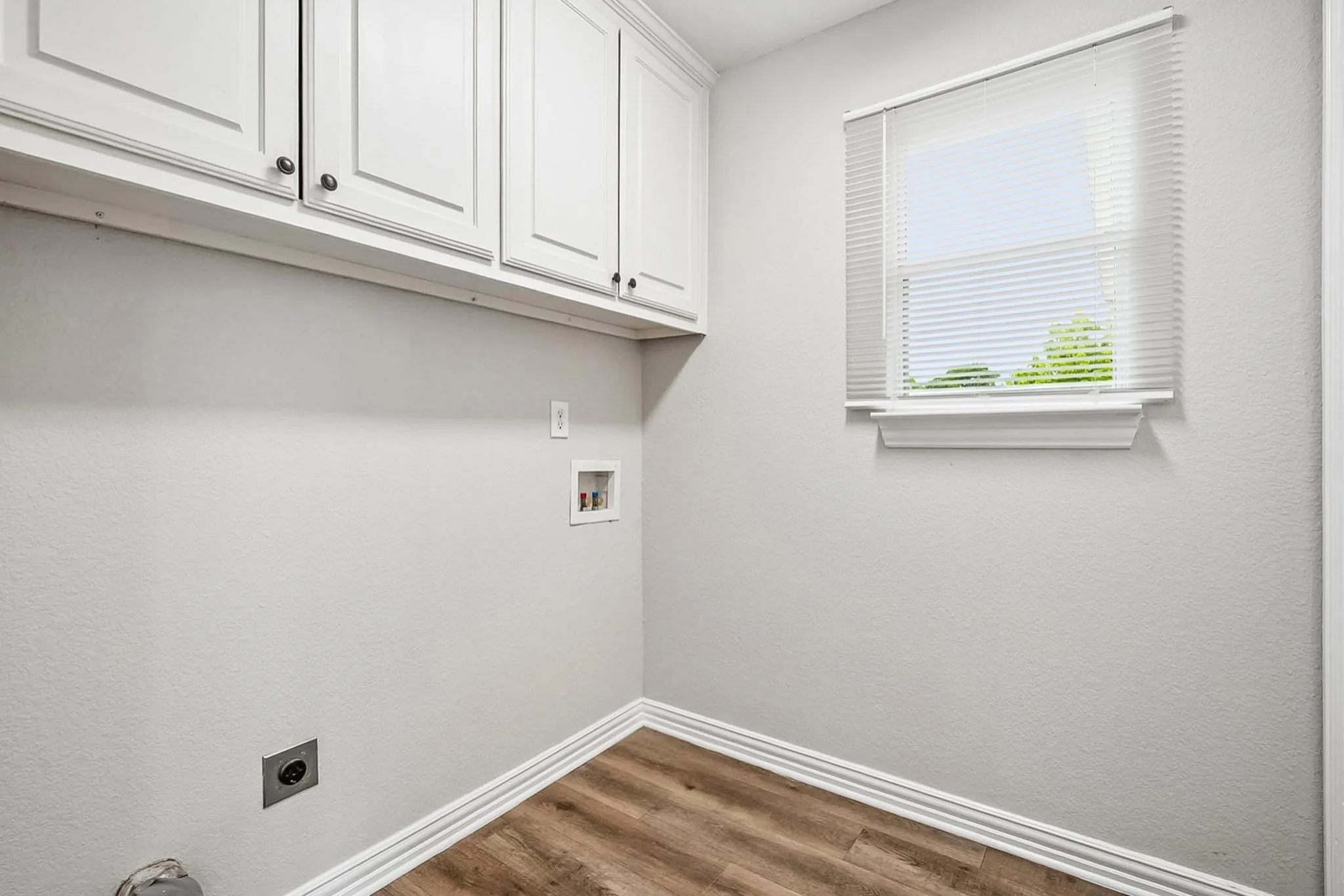 Laundry area featuring wood-finish flooring, white cabinetry with dark hardware, a window with blinds, and white baseboards