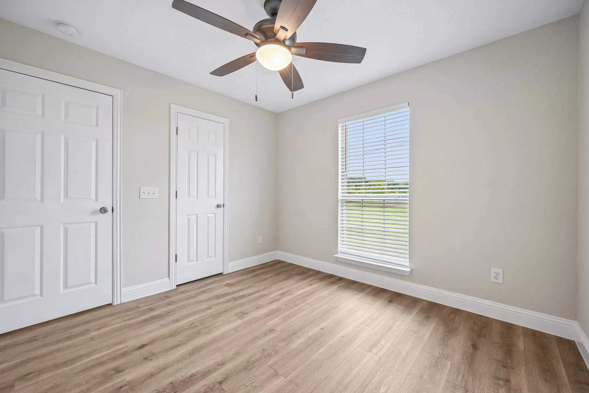 Bright interior space featuring wood-finish flooring, a ceiling fan with integrated lighting, a window with horizontal blinds, and a neutral wall color