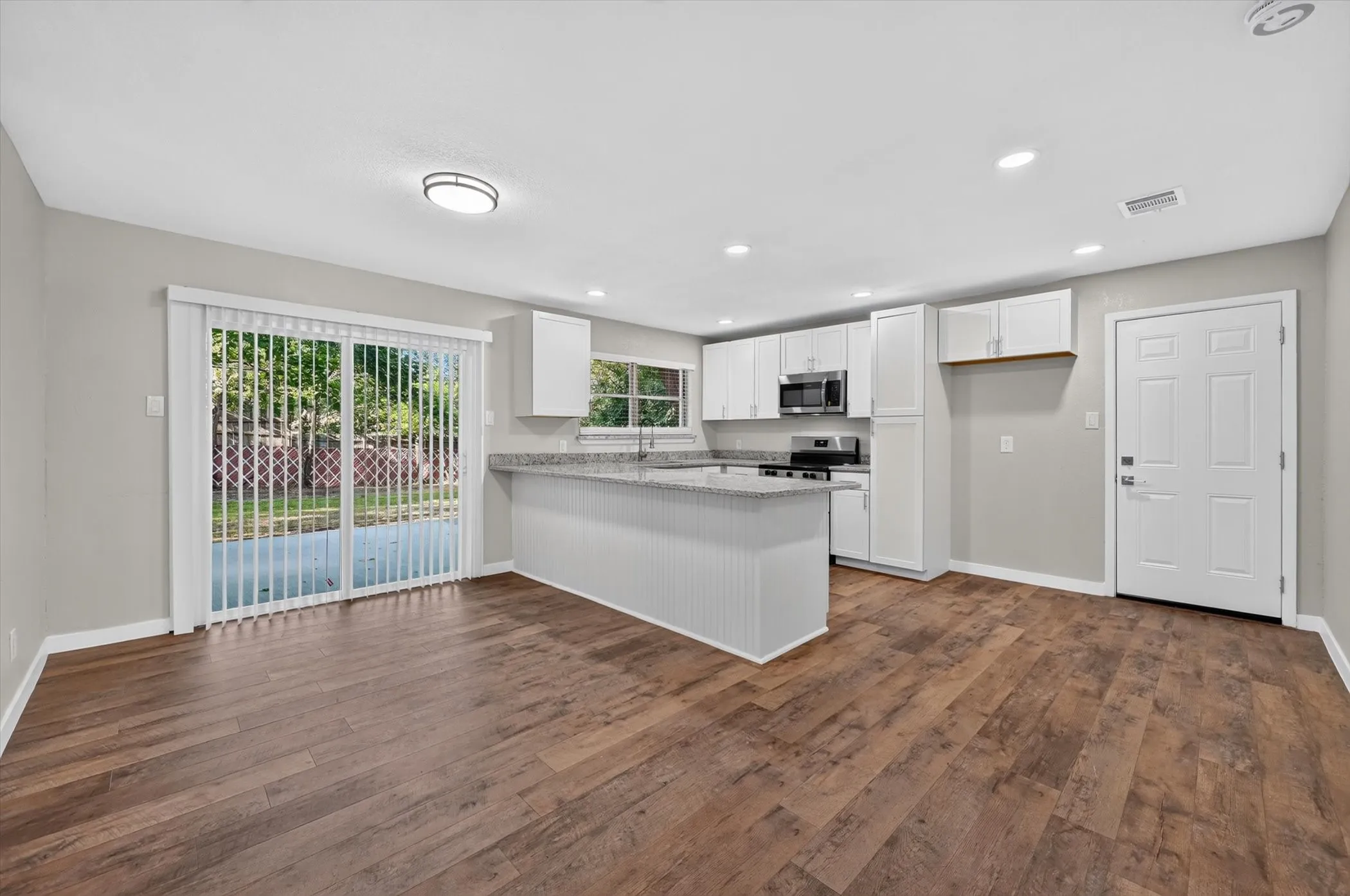 Spacious kitchen and dining area featuring wood-finish flooring, white cabinetry, stainless steel appliances, and a granite-style countertop on the breakfast bar and perimeter