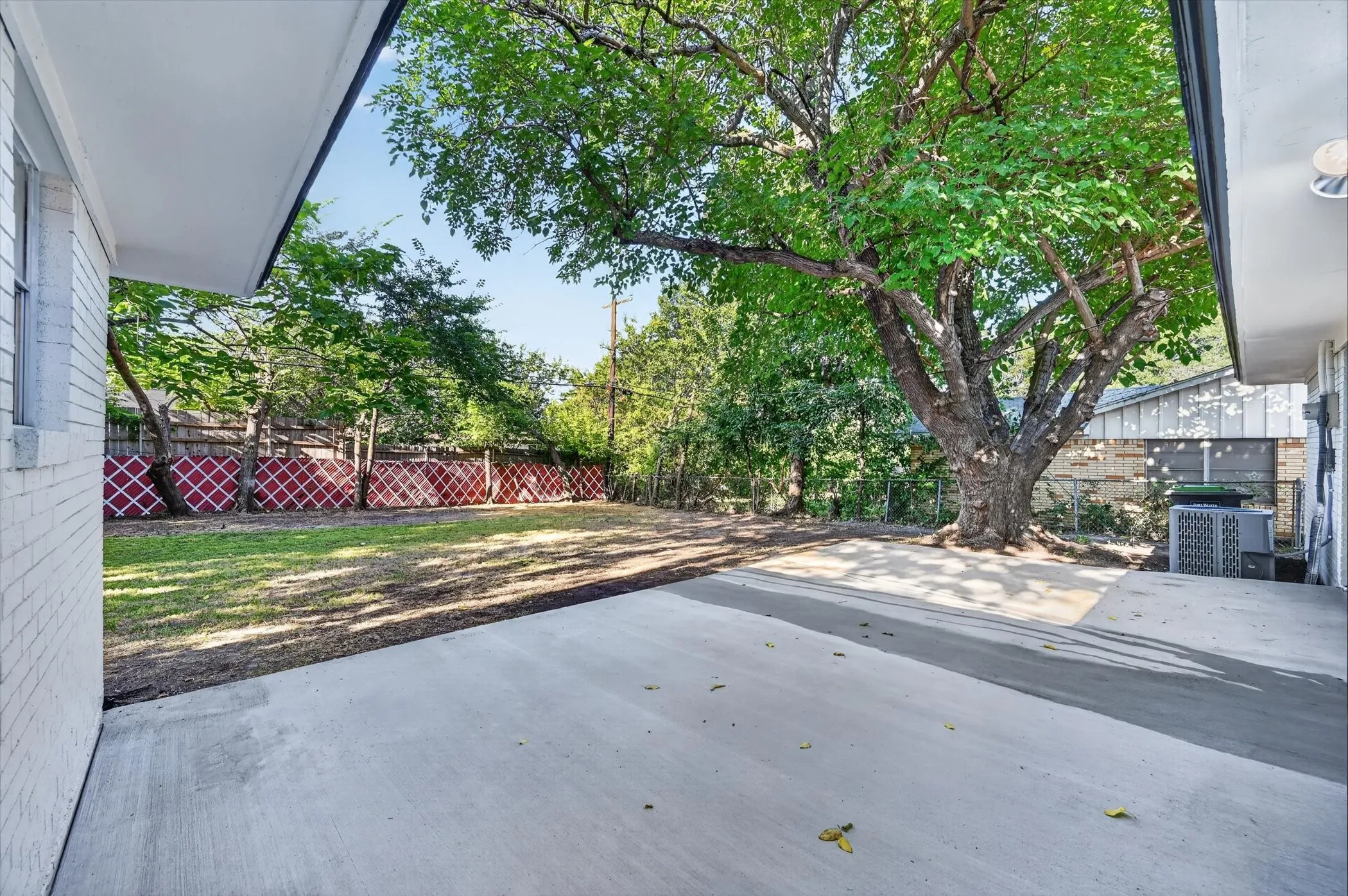 Expansive concrete patio with a mature shade tree, a grass lawn, and a decorative red and white privacy fence