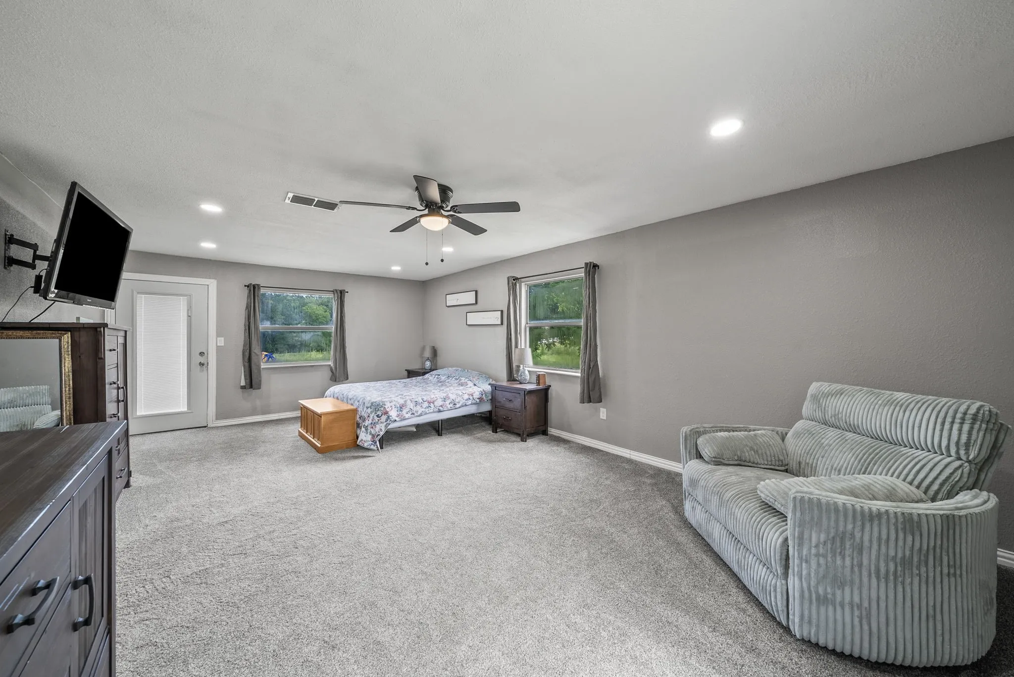 Spacious interior room featuring light gray walls, plush gray carpeting, recessed ceiling lighting, a ceiling fan, and multiple windows