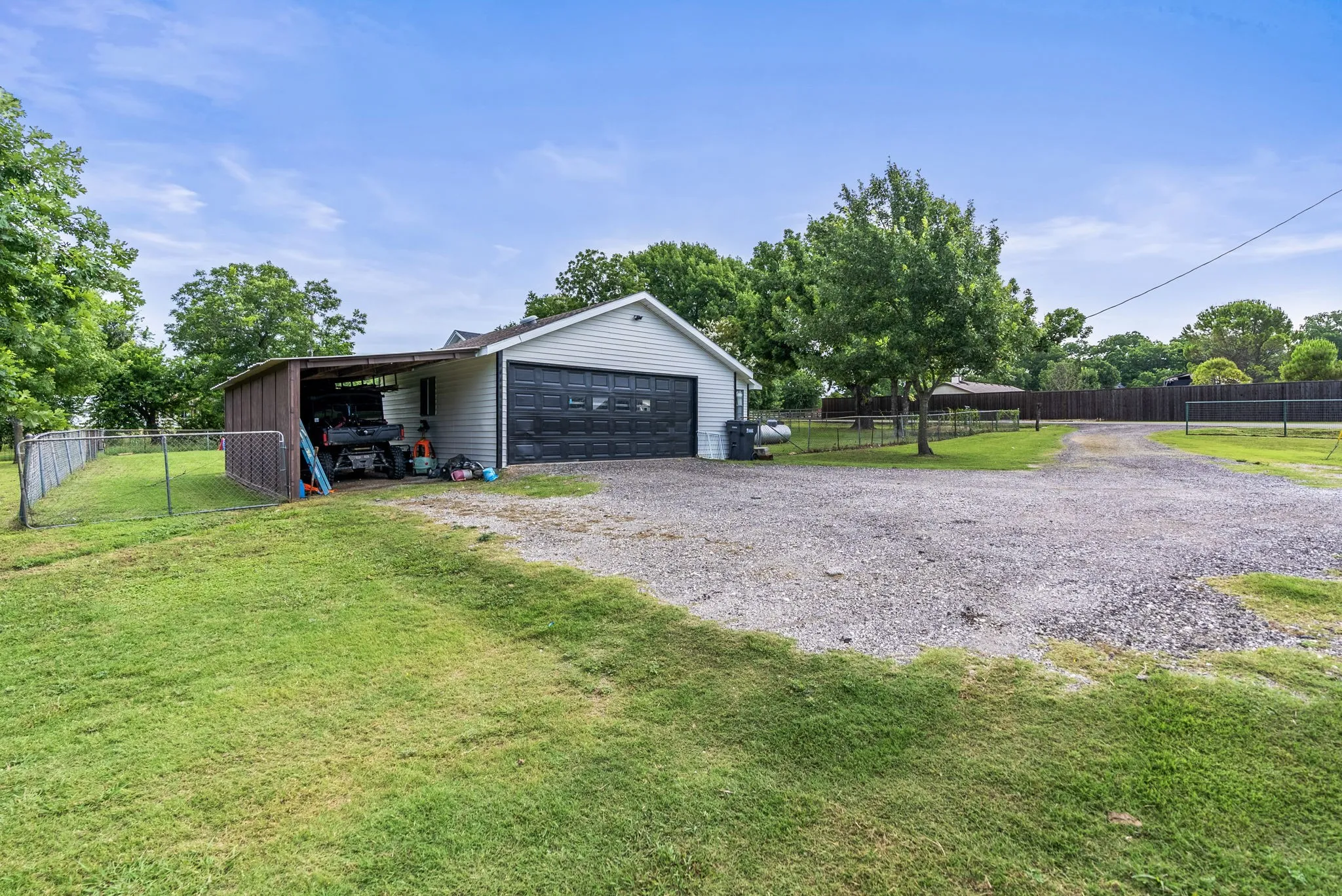 Detached garage with a dark panel garage door, gravel driveway, covered carport, chain-link fencing, and mature trees