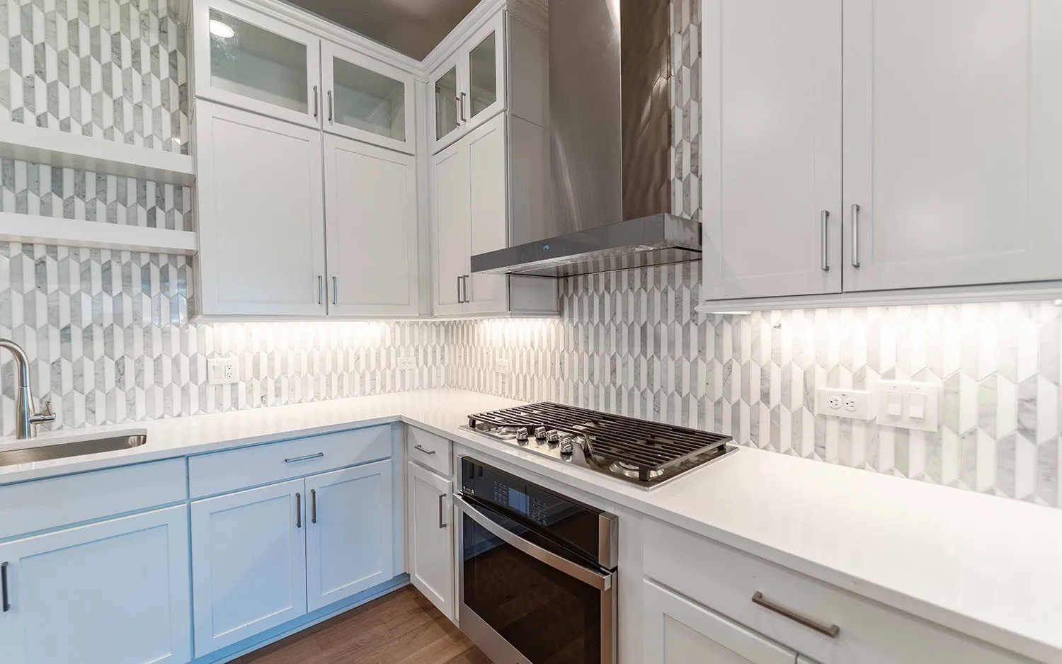Modern kitchen featuring white cabinetry with some upper glass-front cabinets, a white countertop, an integrated stainless steel sink with a gooseneck faucet, a built-in oven, and a gas cooktop with a stainless steel range hood