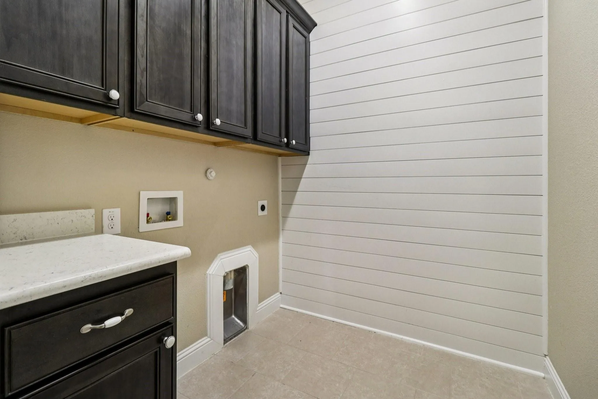 Utility room featuring dark wood cabinetry, white shaker-style shiplap wall treatment, light-toned countertop, and tile flooring