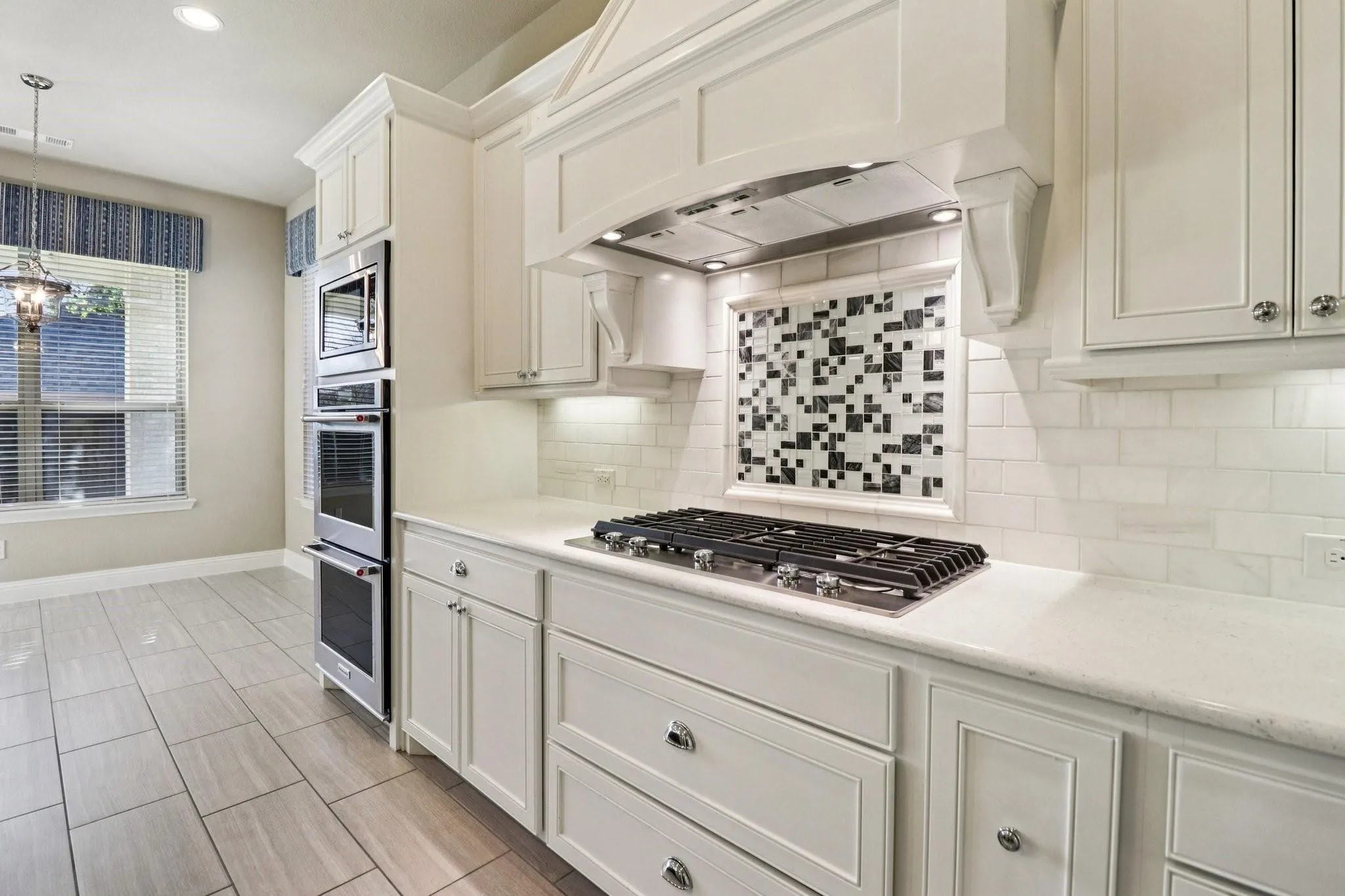 Gourmet kitchen featuring white cabinetry, light-toned countertops, a built-in gas range, a multi-oven tower, and a decorative tile backsplash with a mosaic accent