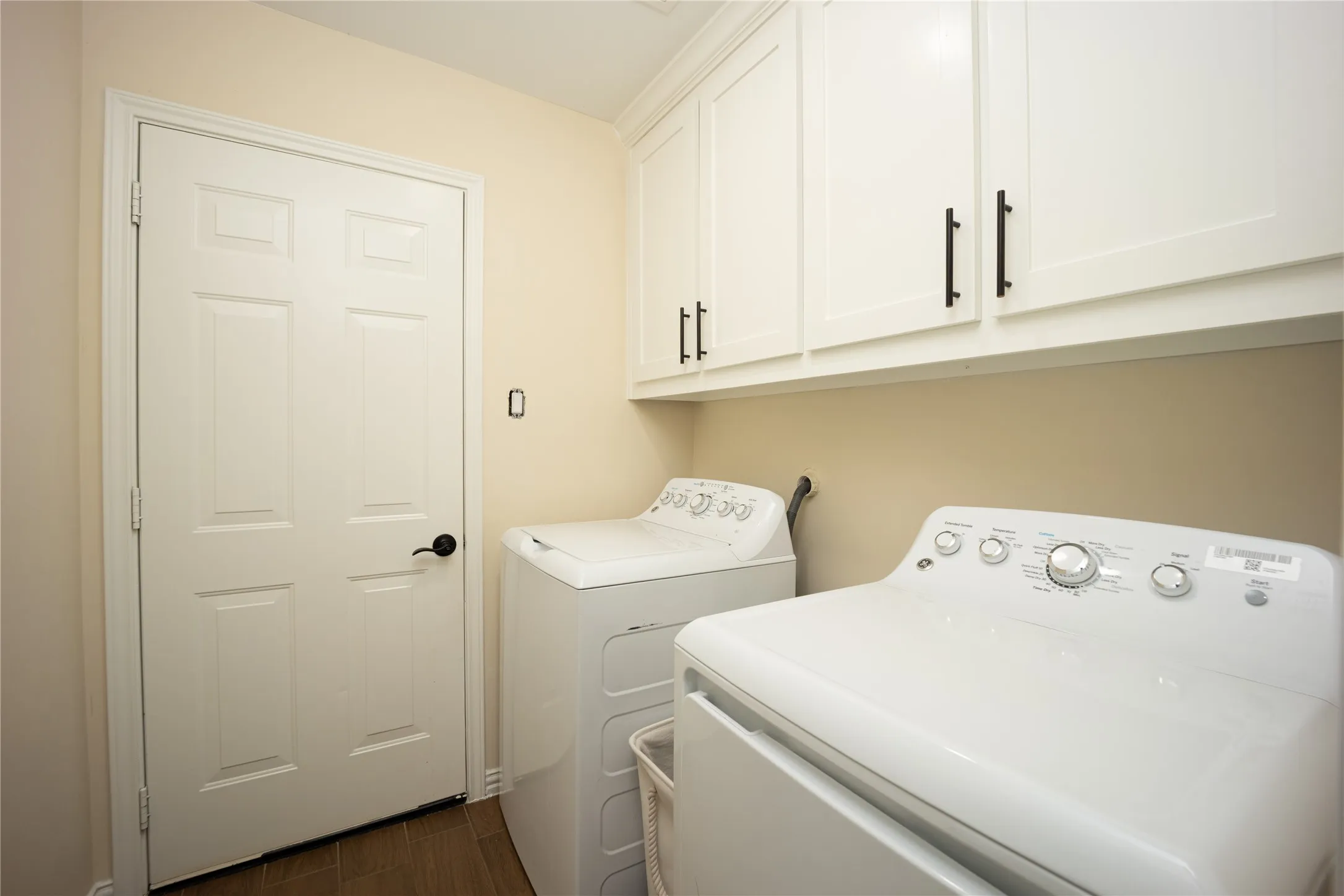 Dedicated laundry space featuring upper white cabinetry with matte black hardware, light neutral wall paint, and wood-finish flooring