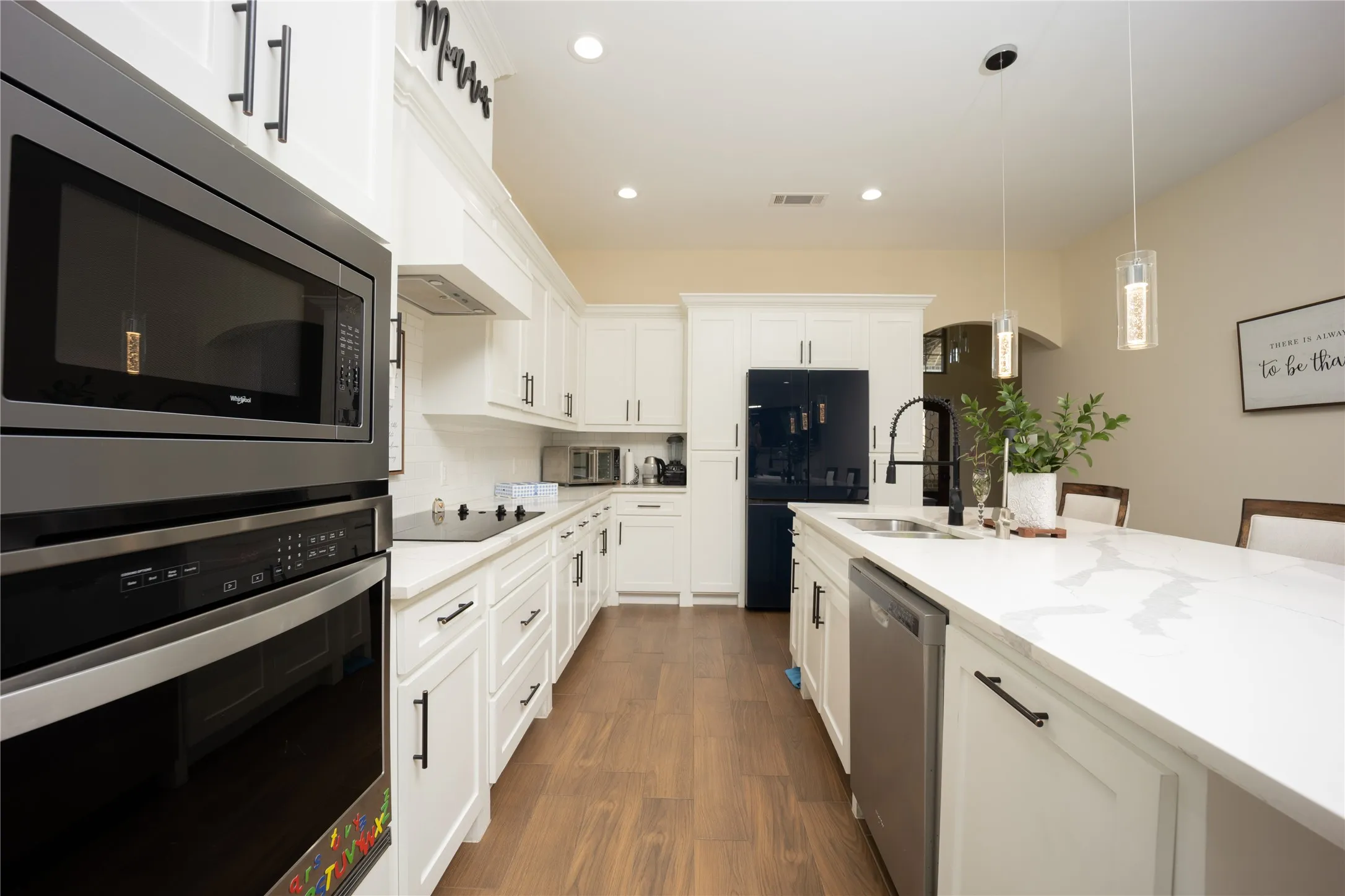 Kitchen featuring white cabinetry with dark hardware, a built-in microwave and oven combination, a smooth cooktop, a dark stainless steel refrigerator, and a kitchen island with a sink and pendant lighting