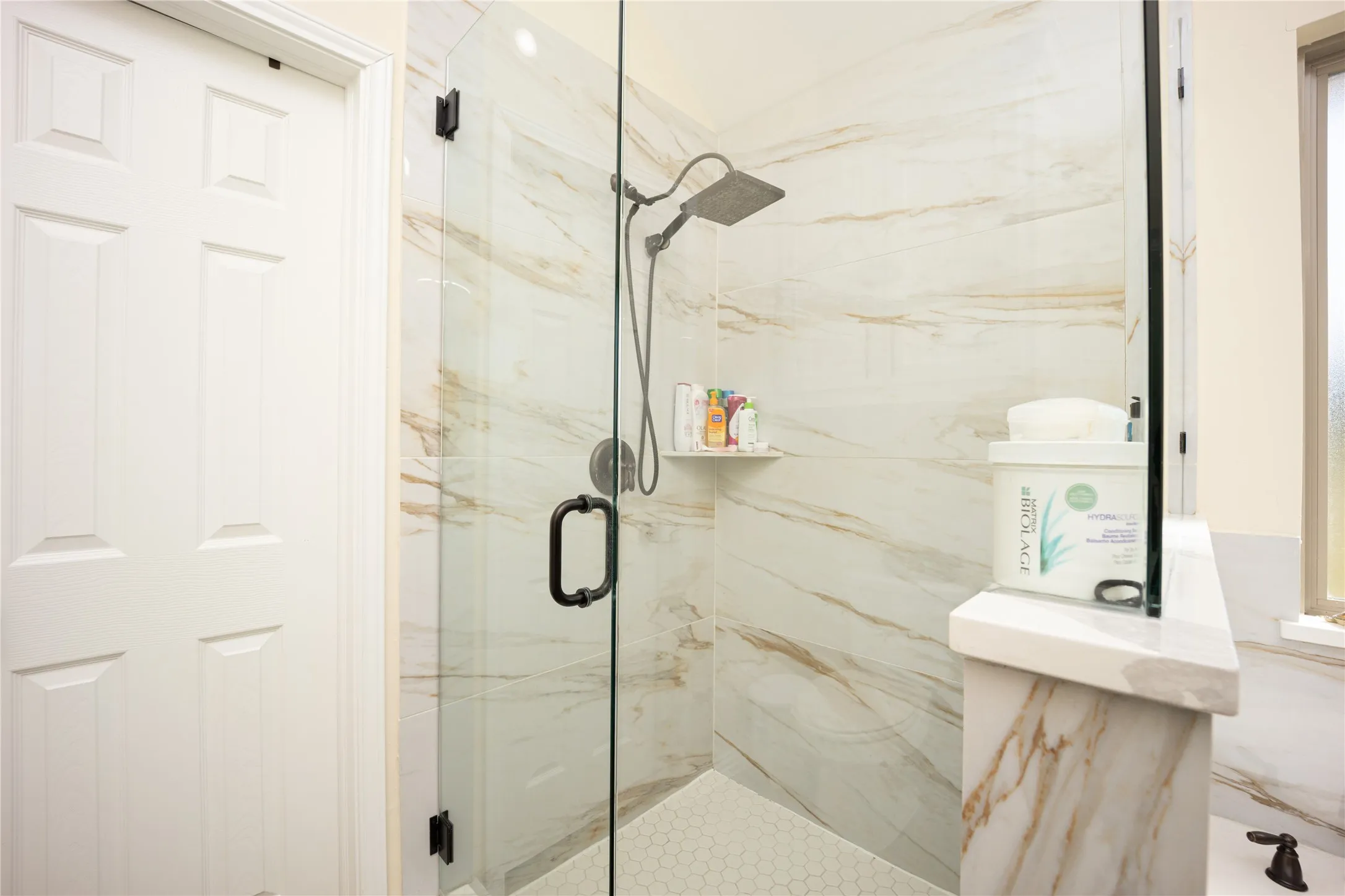 Walk-in shower featuring glass enclosure, large format tile walls with natural stone appearance, rain showerhead, hex tile flooring, and integrated shower niche
