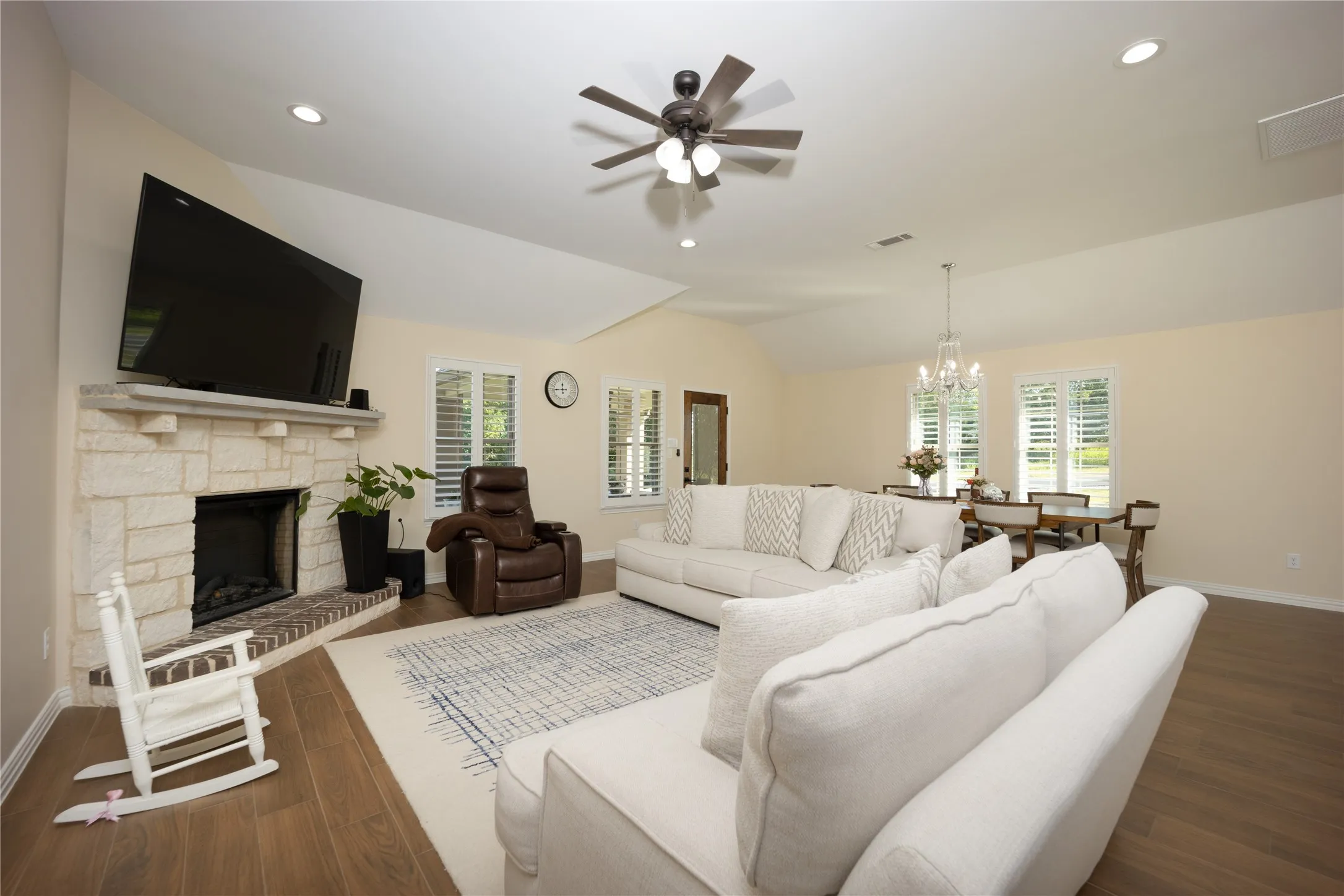 Open-concept living area featuring wood-finish flooring, a stone fireplace with a brick hearth, and vaulted ceilings with recessed lighting