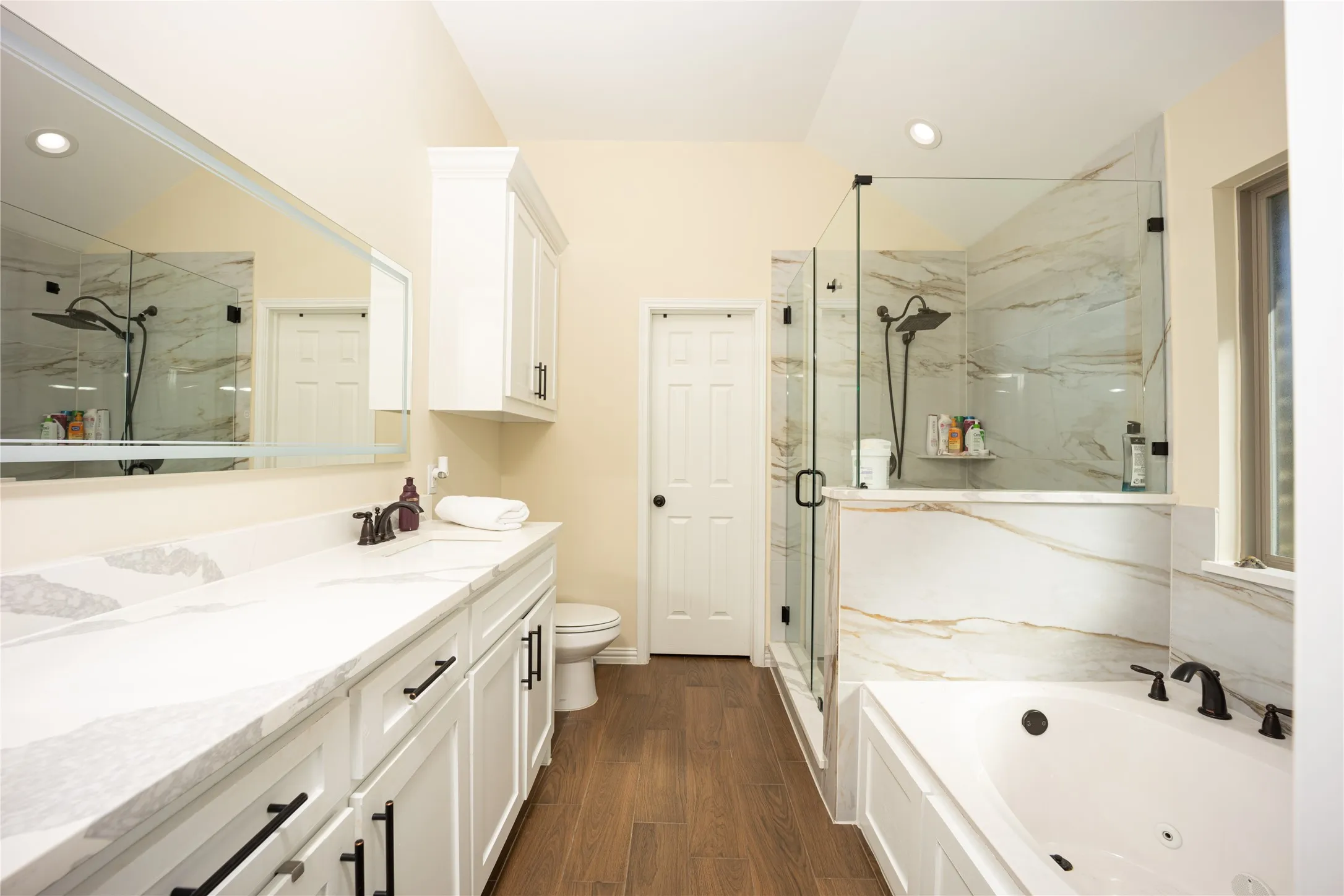 Bathroom featuring a dual vanity with a white countertop and dark hardware, a glass-enclosed shower with tiled walls and a rainfall showerhead, a built-in jetted tub, recessed lighting, and wood-finish flooring