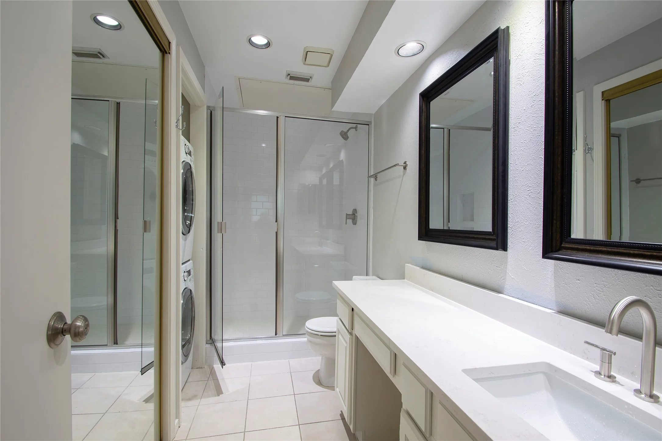White tile flooring in a bathroom featuring a long vanity with an integrated sink and brushed nickel faucet, framed mirrors, a glass-enclosed shower, and recessed lighting