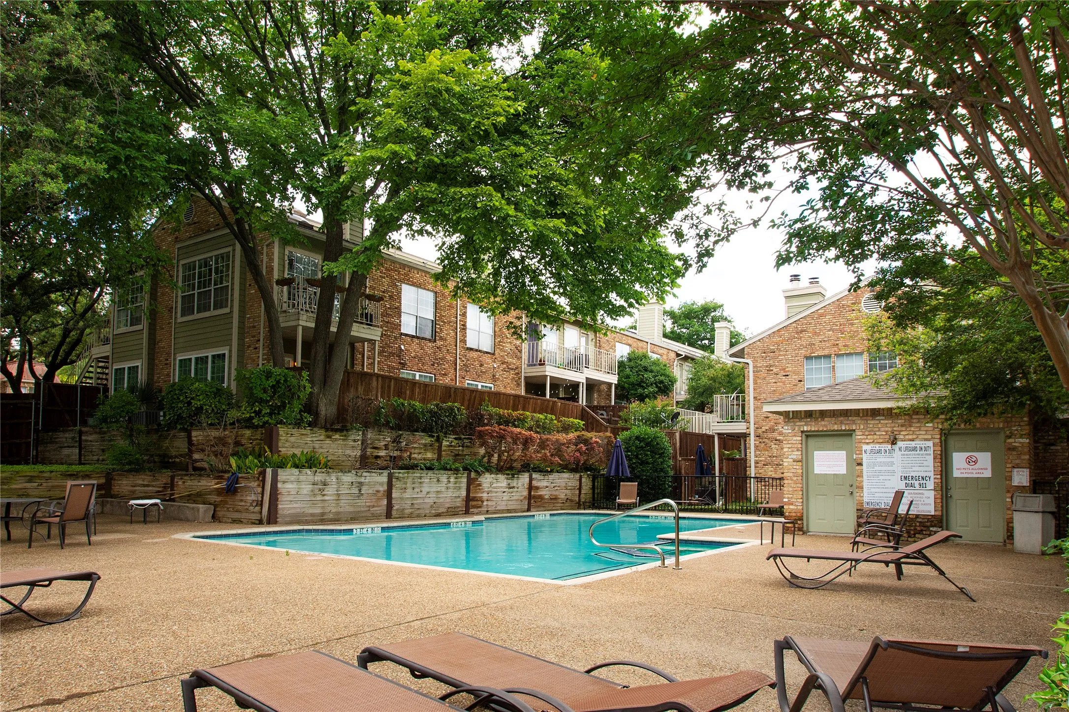 Community swimming pool with a textured concrete deck, surrounded by mature trees, brick-clad buildings, and wooden retaining walls