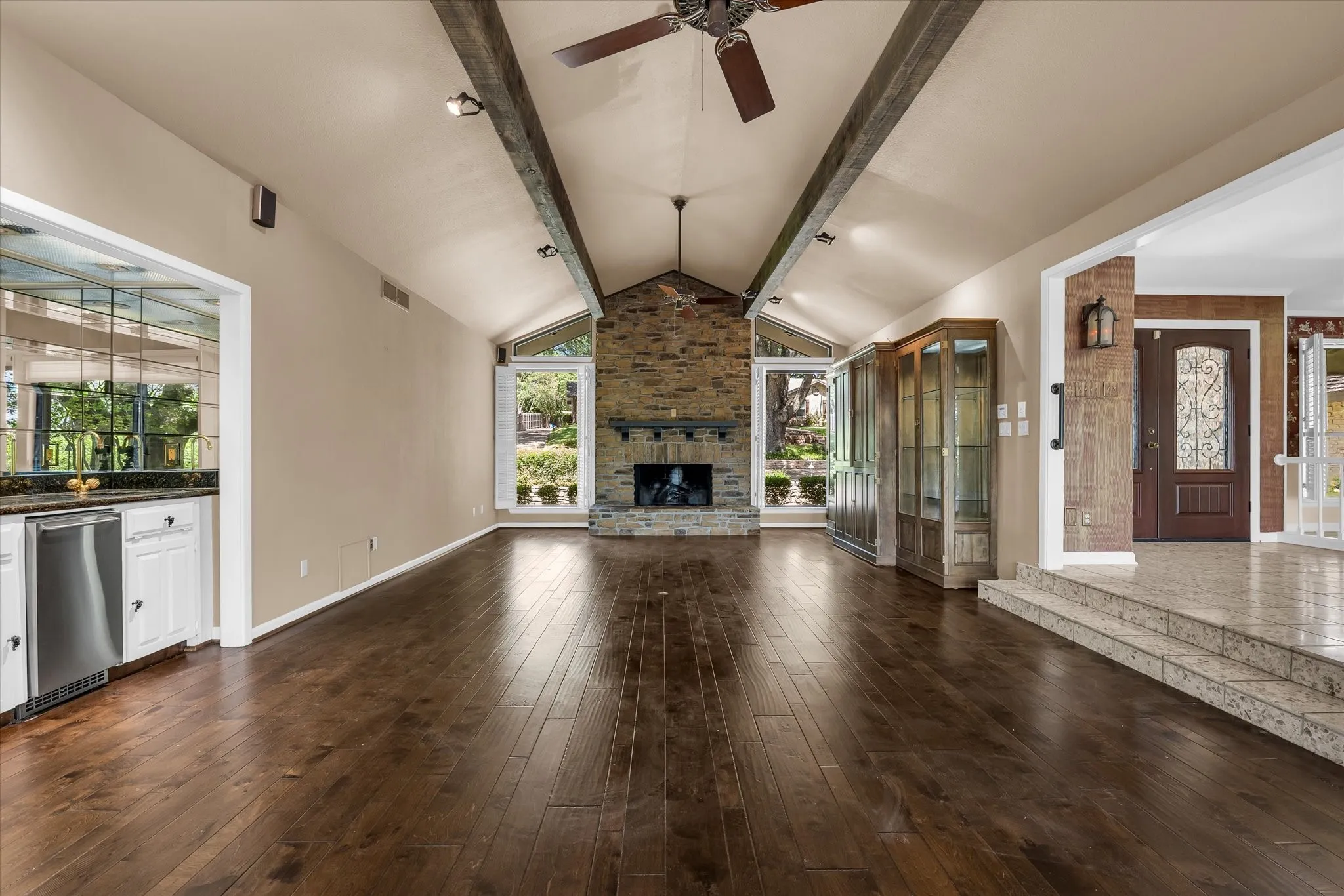 Vaulted ceiling with exposed wood beams, stone-clad fireplace, wood-finish flooring, built-in display cabinetry, and a wet bar with under-counter refrigeration