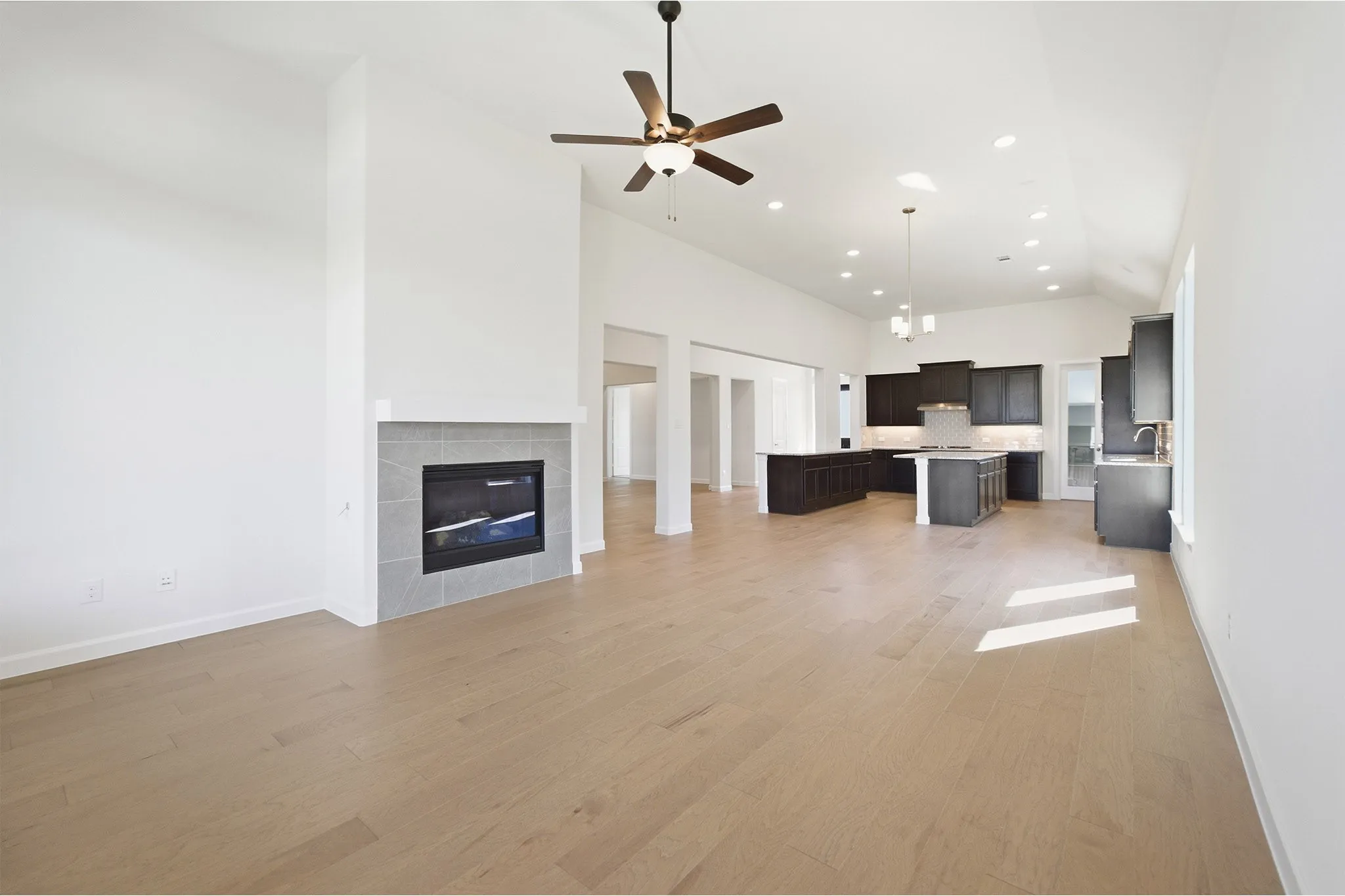 Open-concept living area featuring light wood-finish flooring, a contemporary fireplace with a tile surround, a dark wood-finish ceiling fan, and recessed lighting